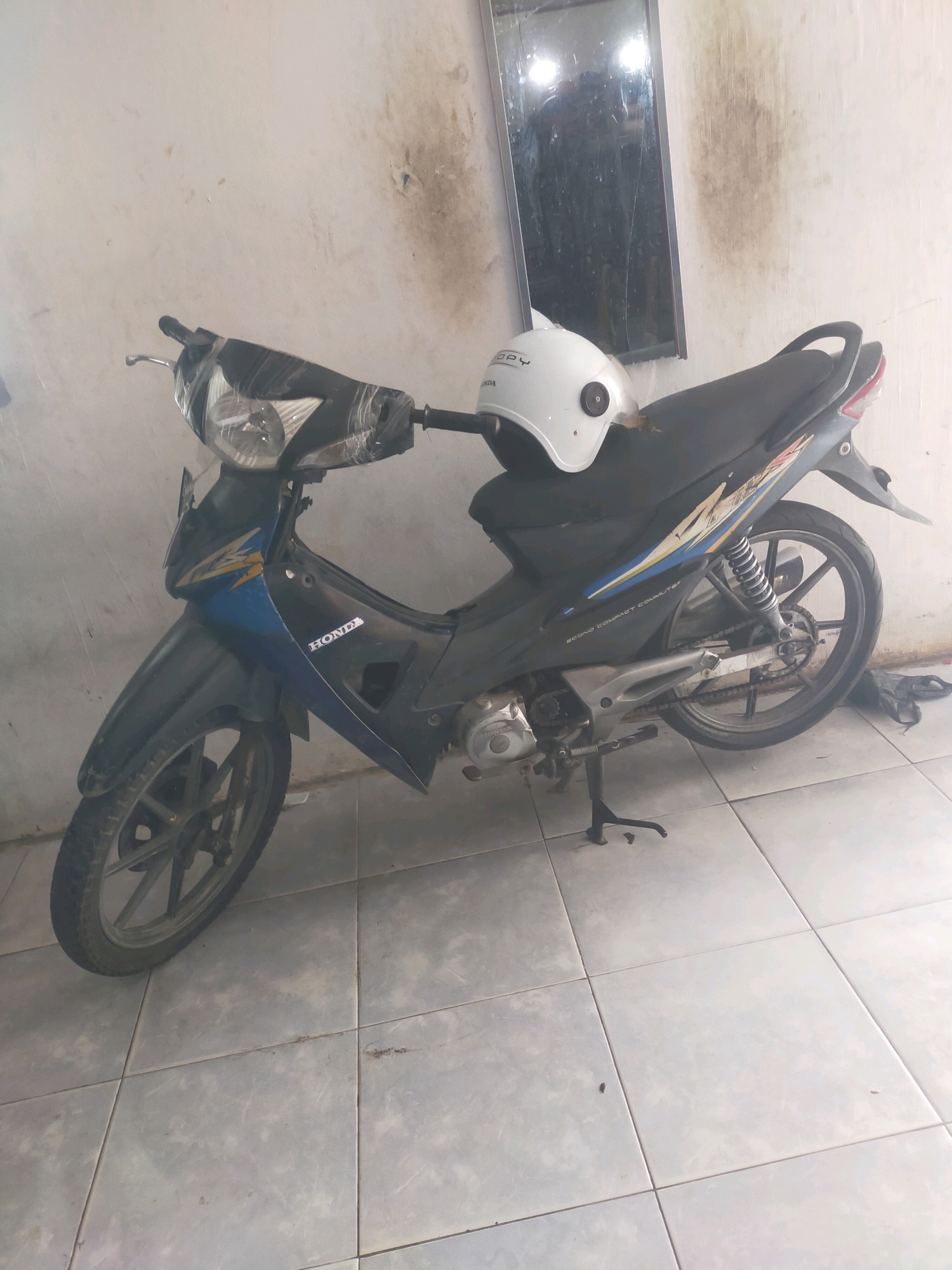 A Honda scooter is parked indoors on a tiled floor. The scooter is black with blue and yellow accents. A white helmet is placed on the seat of the scooter. The walls in the background are white and stained, with a framed mirror hanging on the wall. The scene appears to be in a residential or commercial indoor space in Pekanbaru, Indonesia.