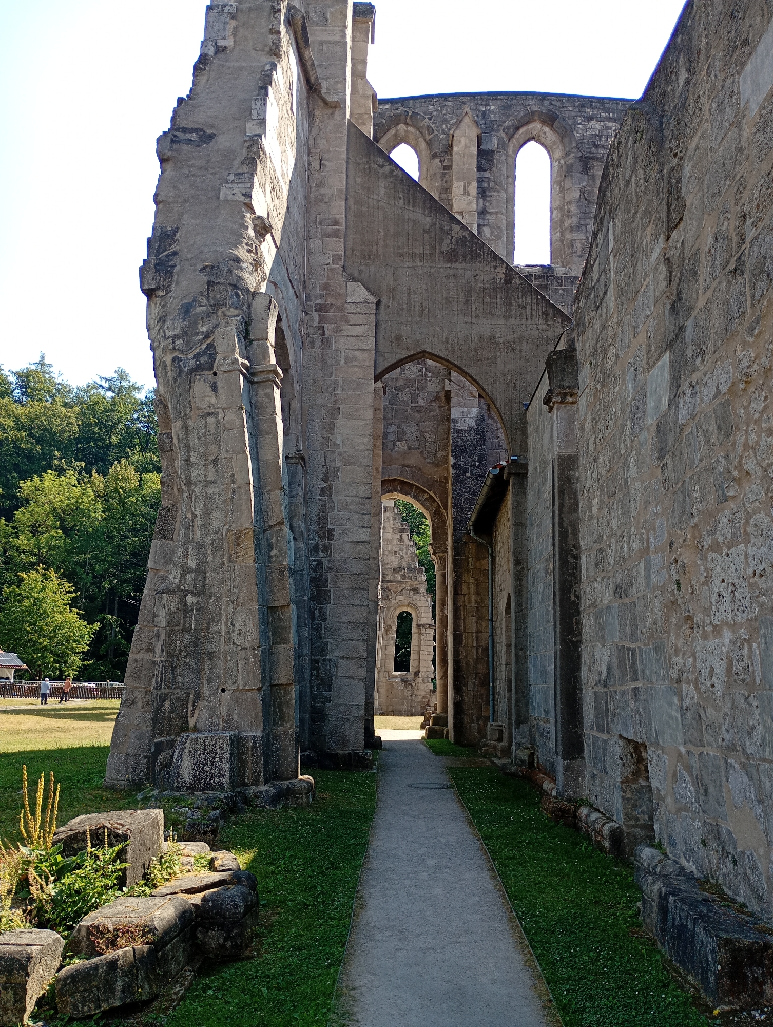 This is a photograph of the ruins of Walkenried Abbey in Walkenried, Germany, taken on a sunny day. The perspective is from a gravel path leading through the stone ruins of the abbey. On the left, a massive, weathered stone wall, part of the abbey's structure, rises dramatically against a bright sky. This wall shows significant damage and erosion, with broken sections and rough textures. Beyond this wall, lush green trees are visible.
The path continues forward, passing under arched openings in the ruined structure. These arches reveal glimpses of further ruined walls and windows in the background, also made of stone. To the right of the path, a thick, stone-built wall of the abbey runs parallel, constructed from large, unevenly shaped blocks. Some sections of this wall also appear weathered and are partially obscured by shadows.
The ground flanking the path is covered in green grass, with some patches of weeds and a few large, broken stone blocks in the foreground on the left. In the distance, beyond a wooden fence, a grassy field with a few scattered trees and two people walking can be seen. The overall impression is one of ancient grandeur and decay, with the bright sunlight highlighting the textures and forms of the stone architecture and the surrounding vegetation.