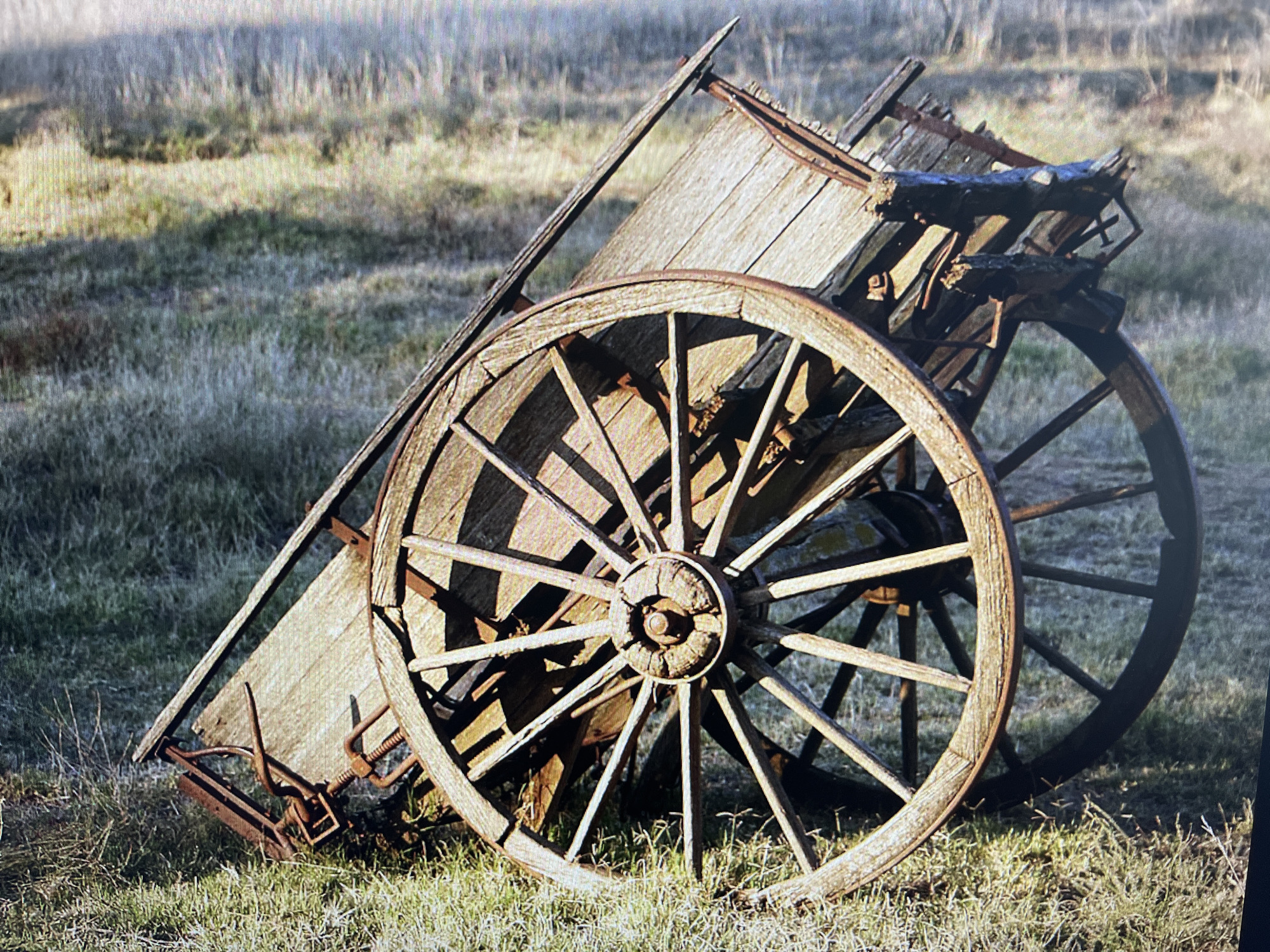 The image captures an old, weathered wooden wagon or cart, seemingly abandoned in a grassy field in Varna, Bulgaria. The wagon is the central subject, positioned at an angle, with its large spoked wheels prominently featured. The wood of the wagon is aged and rustic, showing signs of decay, wear, and exposure to the elements, with some planks appearing broken or missing. Metal components, such as the wheel rims and structural supports, exhibit significant rust. The wagon appears to be leaning slightly to one side.
The setting is a natural, open area covered in dry, green-brown grass. The lighting suggests either late afternoon or early morning, characterized by a golden hue on parts of the grass and the wagon, and discernible shadows indicating a low sun angle. No activity is taking place; the wagon is stationary and evokes a sense of history or abandonment. There are no people visible in the scene.
A notable technical detail of the image itself is a visible grid-like pattern or pixelation across the entire frame, particularly noticeable in the background, which might suggest it is a photograph taken from a screen or a low-resolution digital capture. There is no visible text anywhere in the image.