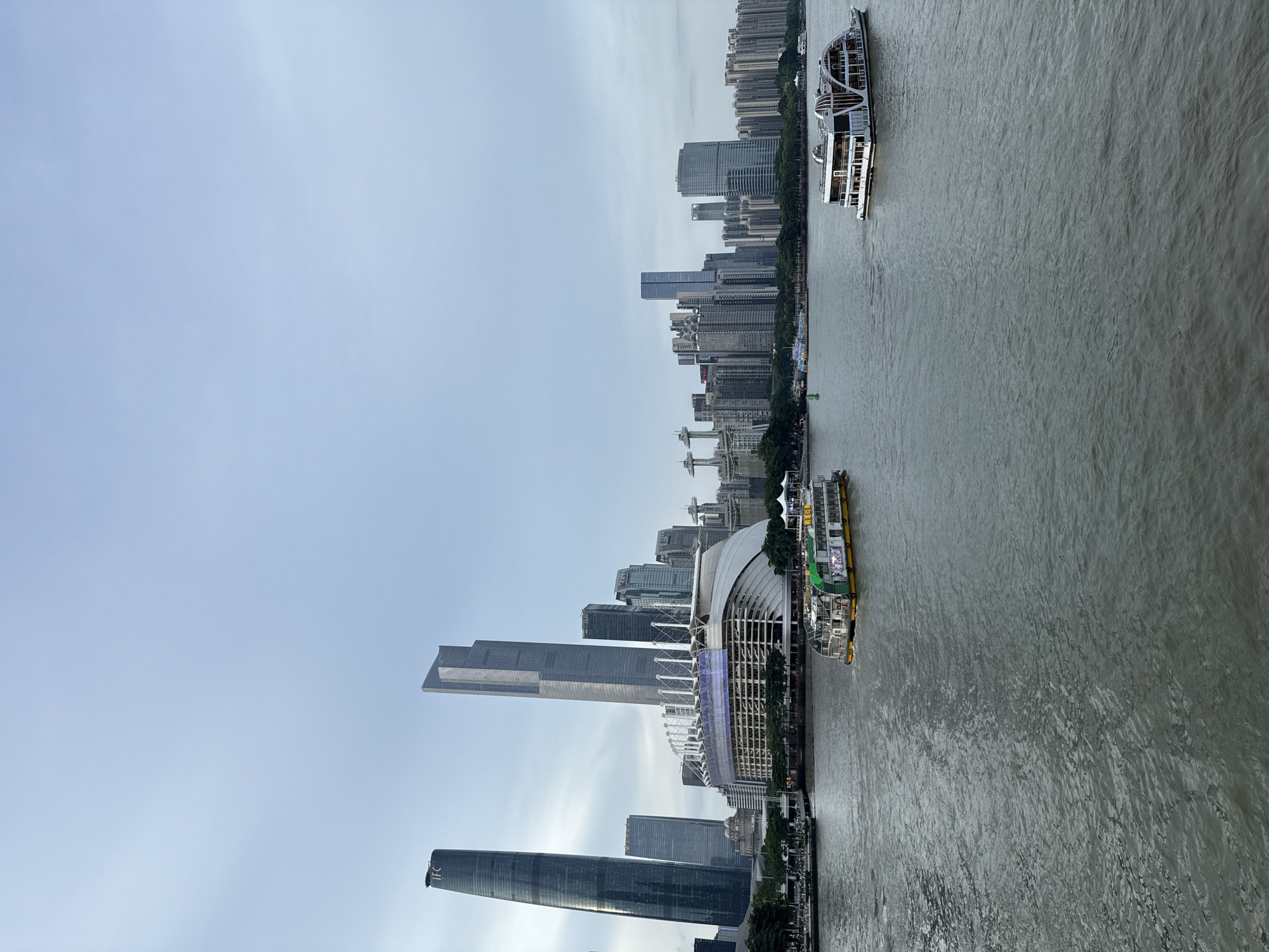 The image captures a wide river flowing through a modern city skyline, likely in Haizhu District, China. The sky is overcast with a pale blue hue. On the left, two prominent skyscrapers dominate the view, the taller one on the far left bearing the "IFC" logo. Adjacent to these, a large, curved white structure with numerous white supports, possibly a stadium or exhibition center, is visible. The river is populated by several vessels. In the foreground, a long, multi-decked white boat with arched windows and a distinctive paddlewheel-like stern cruises to the right. Further back, closer to the shore, another boat with a green and white superstructure is moving left. The riverbanks are lined with trees and a bustling promenade, with numerous buildings of varying heights forming the urban landscape. The water exhibits a textured surface with ripples and waves, reflecting the subdued light.