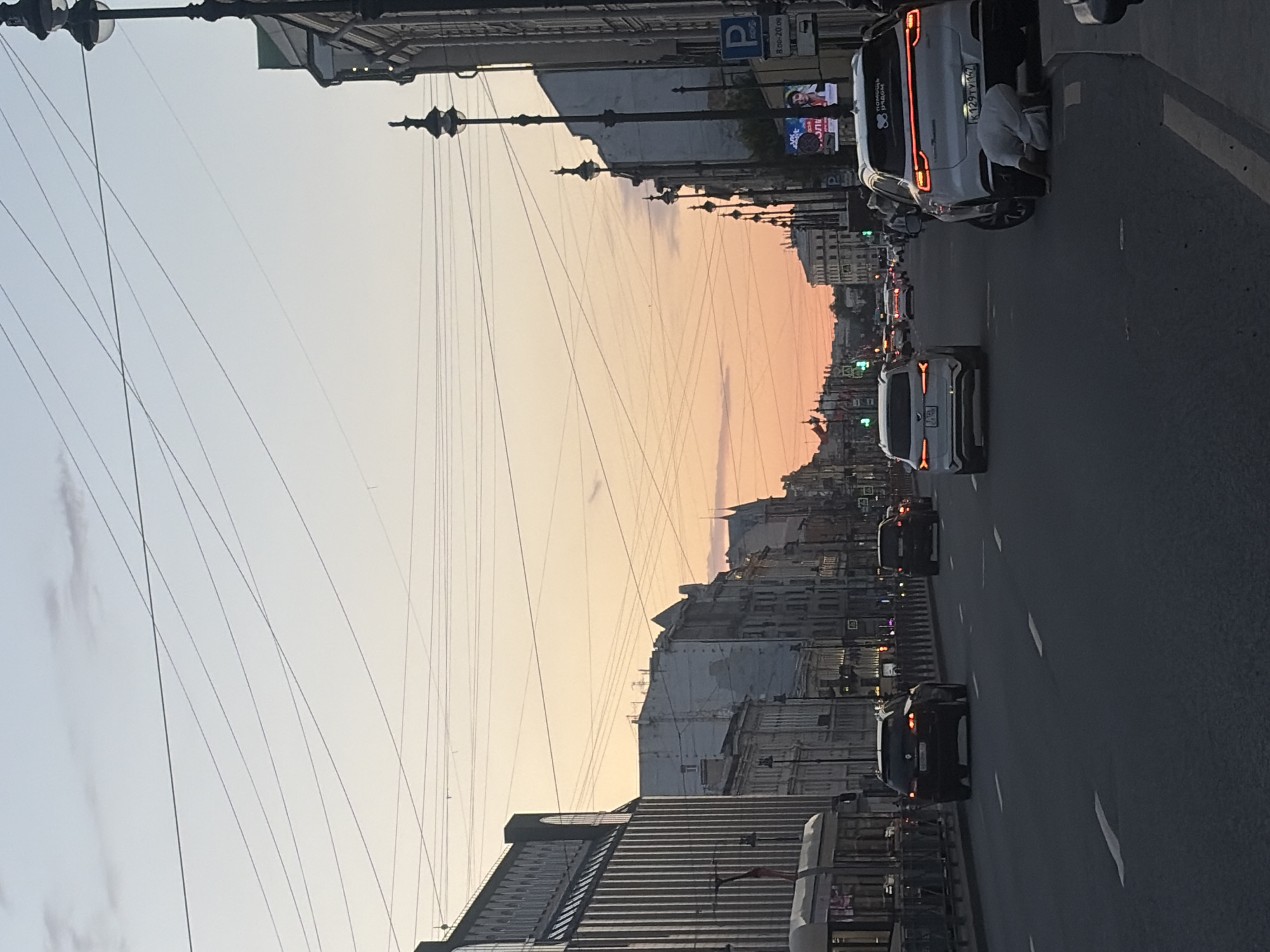 The image depicts a street scene in Saint Petersburg, Russia, during twilight. The sky is a gradient of soft orange and pale blue, with numerous overhead wires crisscrossing the frame. On the left, a modern building with a ribbed facade and arched windows stands next to older, multi-story buildings with traditional architecture. A tram or streetcar is visible on the far left.
The street itself is busy with traffic. Several cars are driving on the road, including a white SUV in the foreground on the right, which has a person in white clothing kneeling beside its rear tire. Other vehicles are seen further down the street. Streetlights with ornate designs line the right side of the road, and a "P" sign indicating parking is visible on a pole. Billboards with advertisements are also present on the buildings. The overall impression is of an urban environment at the end of the day, with both modern and historic elements coexisting.