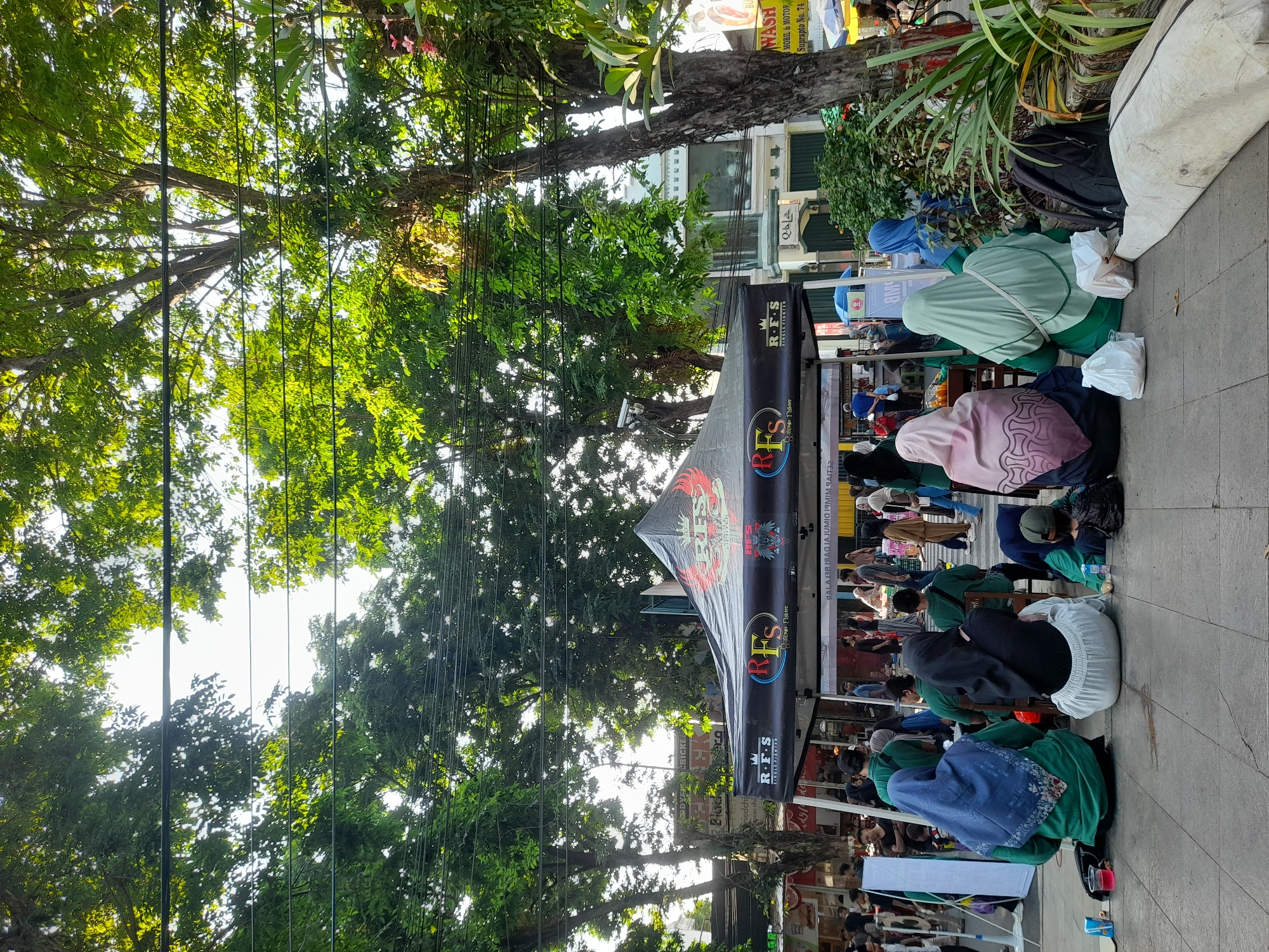 The image captures an outdoor scene in Purwodadi, Indonesia, featuring a black tent with a logo that appears to read "RFS SINGLE FIGHTER" multiple times. Several people are gathered under and around the tent, many of whom are women wearing hijabs in various colors, including green, black, pink, and blue. Some are sitting on the ground, while others are standing or walking. The tent is set up on a paved area, possibly a sidewalk or plaza, with lush green trees providing shade overhead. In the background, there are buildings, other tents or stalls, and more people. The overall atmosphere suggests a public event or market.