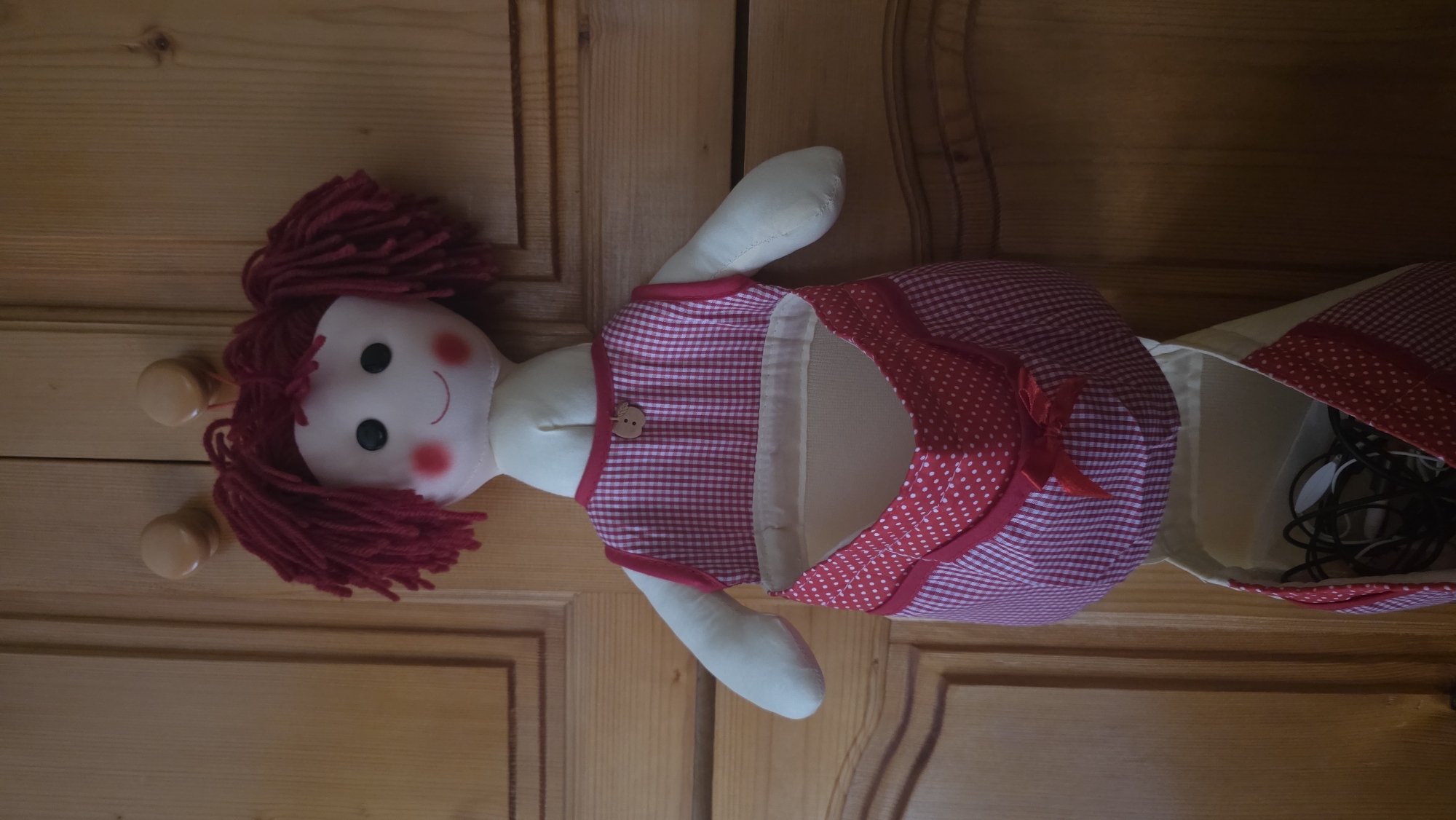 A fabric doll, serving as a storage bag or holder, is depicted hanging against a wooden background in an indoor setting in Blagoevgrad, Bulgaria. The image is oriented sideways, presenting the doll horizontally.
The doll features a cream-colored head with red yarn hair styled into pigtails. Its face has black button eyes, a simple red stitched smile, and rosy red blush on its cheeks. Two light brown wooden pegs or hooks are visible near the top of the doll's head, indicating a possible hanging mechanism.
The doll's body is dressed in a red and white checkered fabric outfit, resembling a pinafore or dress. The upper part of the garment has narrow red trim, and a small, light brown wooden button shaped like an apple is attached to the chest. The lower section of the dress opens up to form a functional pouch or bag. This internal compartment is lined with cream-colored fabric and trimmed with red polka dot fabric, and it contains a cluster of tangled dark and light-colored cables or wires.
The background is a light brown wooden surface, possibly a door or wall paneling, with visible wood grain and recessed details, contributing to a cozy, domestic atmosphere. The doll is presented passively in its role as a storage item. There is no visible text in the image.
