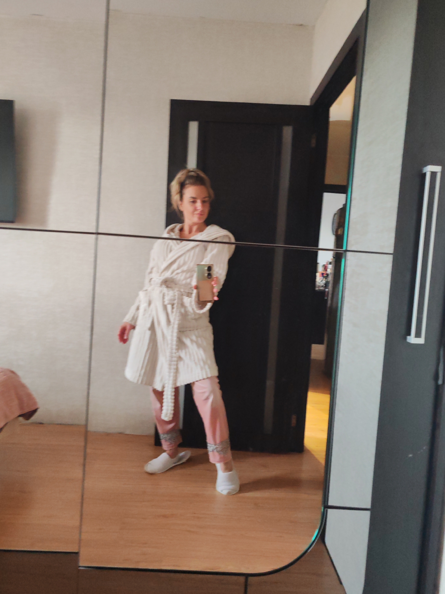 The image displays a mirror selfie of a woman inside what appears to be a bedroom or dressing area, likely in Petropavlovsk-Kamchatsky, Russia. The woman, with blonde hair tied up, is seen from a slight angle, reflected in a large mirror or mirrored closet doors.
She is wearing a light-colored, possibly off-white or cream, hooded bathrobe with a textured, striped pattern, cinched at the waist with a matching tie belt. Beneath the robe, she has on light pink pajama-style pants with decorative grey or silver detailing near the ankles. On her feet, she wears white slippers or socks. In her right hand, she holds a light-colored smartphone, presumably taking the picture, with its camera bump clearly visible. Her expression is neutral to slightly smiling.
The reflected background reveals elements of a modern indoor space. The walls are a light, neutral color with a subtle texture. A dark brown or black doorframe and door are prominent behind her, leading into another room that appears to have a warm-toned wooden floor. To the left side of the mirror, a small section of a bed with a pink or peach-colored blanket is visible. To the right, a dark vertical panel, possibly part of a wardrobe, features a silver bar-style handle. The lighting suggests it is daytime. No discernible text is visible within the image.
