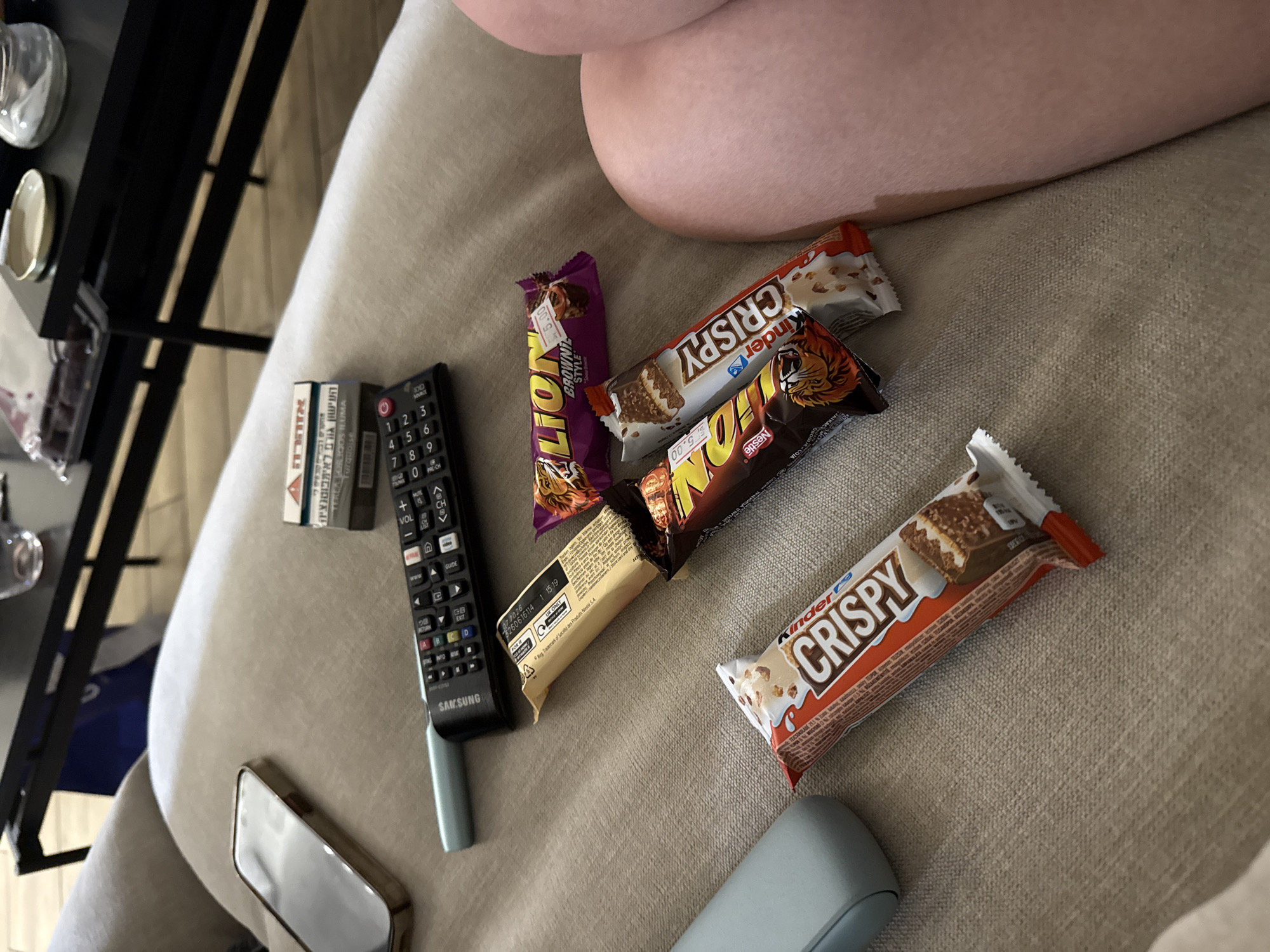 The image depicts an indoor scene focused on a beige fabric couch, where a person is relaxing, partially shown by their bare leg or thigh in the upper right.
Scattered on the couch are various items. Several candy bars are present, including two "Lion" bars (one explicitly labeled "Brownie Style") and two "Kinder Crispy" bars. Many of the wrappers are opened, suggesting consumption. A black Samsung TV remote control lies near the center, alongside a small pack of "REAL GLORIOUS ILUMA Turquoise" cigarettes. The cigarette pack features a Hebrew health warning: "העישון גורם לאימפוטנציה" (Smoking causes impotence).
Further details include a partially visible smartphone with a golden frame and a light blue/grey object, possibly a portable speaker or earbud case, both resting on the couch. In the blurred background to the left, a black metal shelving unit or table holds reflective objects like glasses or small dishes.
Visible text on the remote includes "SAMSUNG," numeric buttons, and function keys like "VOL," "CH," "GUIDE," "NETFLIX," and "WWW." The candy wrappers display "LION," "Kinder," "CRISPY," and "Nestlé." A price sticker "₪5.00" (5 Israeli Shekels) is visible on one Lion bar, along with an expiry date "17.2026," "UK ONLY," and "® Reg. Trademark of Société des Produits Nestlé S.A."
The scene conveys a casual atmosphere of relaxation and snacking, possibly while watching television, set in a home environment. The Hebrew text on the cigarette pack provides a geographical context pointing to Israel.