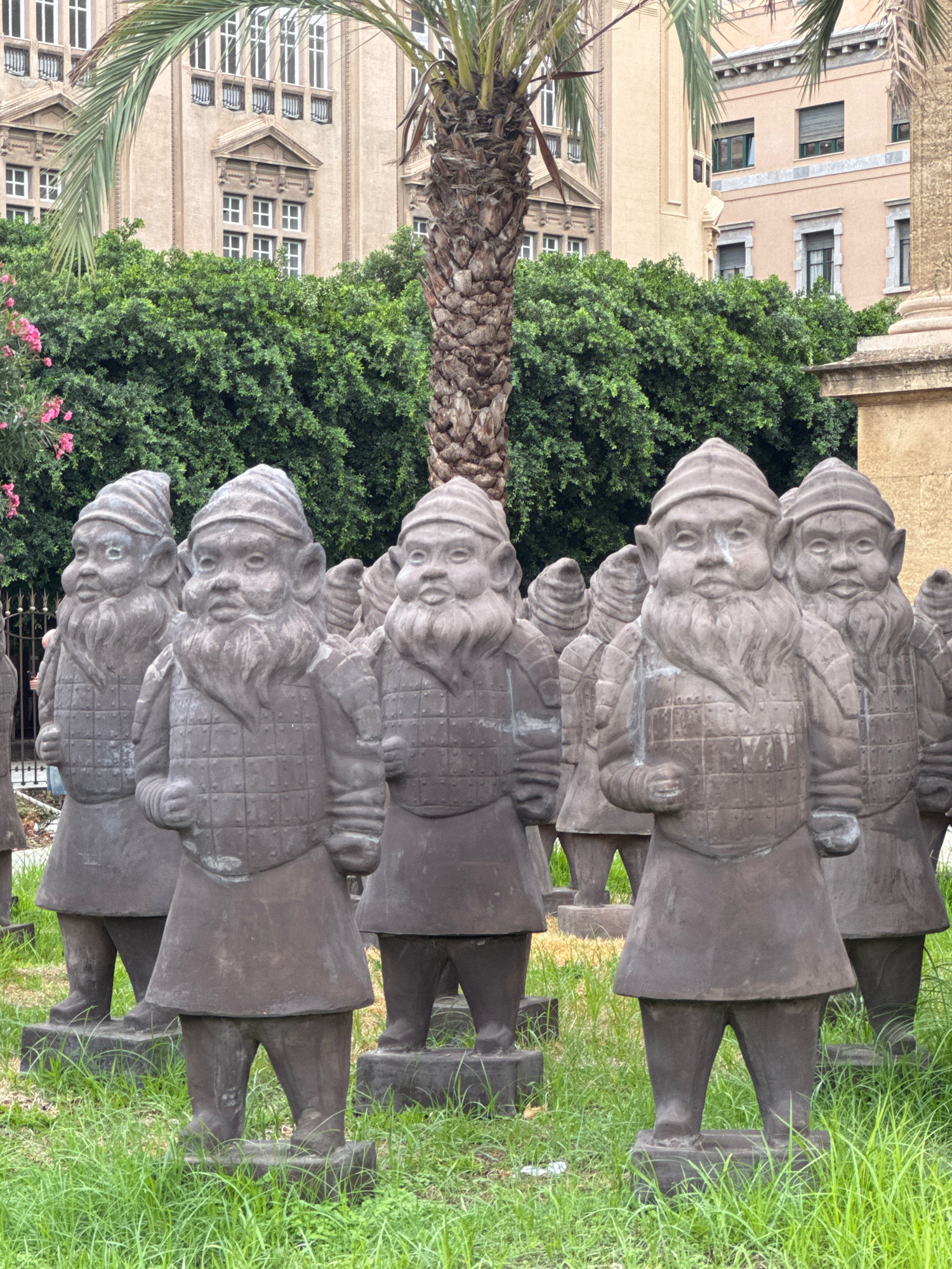A group of stone gnome statues, resembling Terracotta Army figures, are displayed outdoors in a grassy area in Palermo, Italy. The statues are arranged in rows, facing forward, and each is depicted wearing armor and a hat. Behind the statues, lush green trees and foliage form a backdrop, with a tall palm tree prominently featured in the center. In the background, a pale building with multiple windows and architectural details suggests an urban setting. The lighting indicates it is daytime.