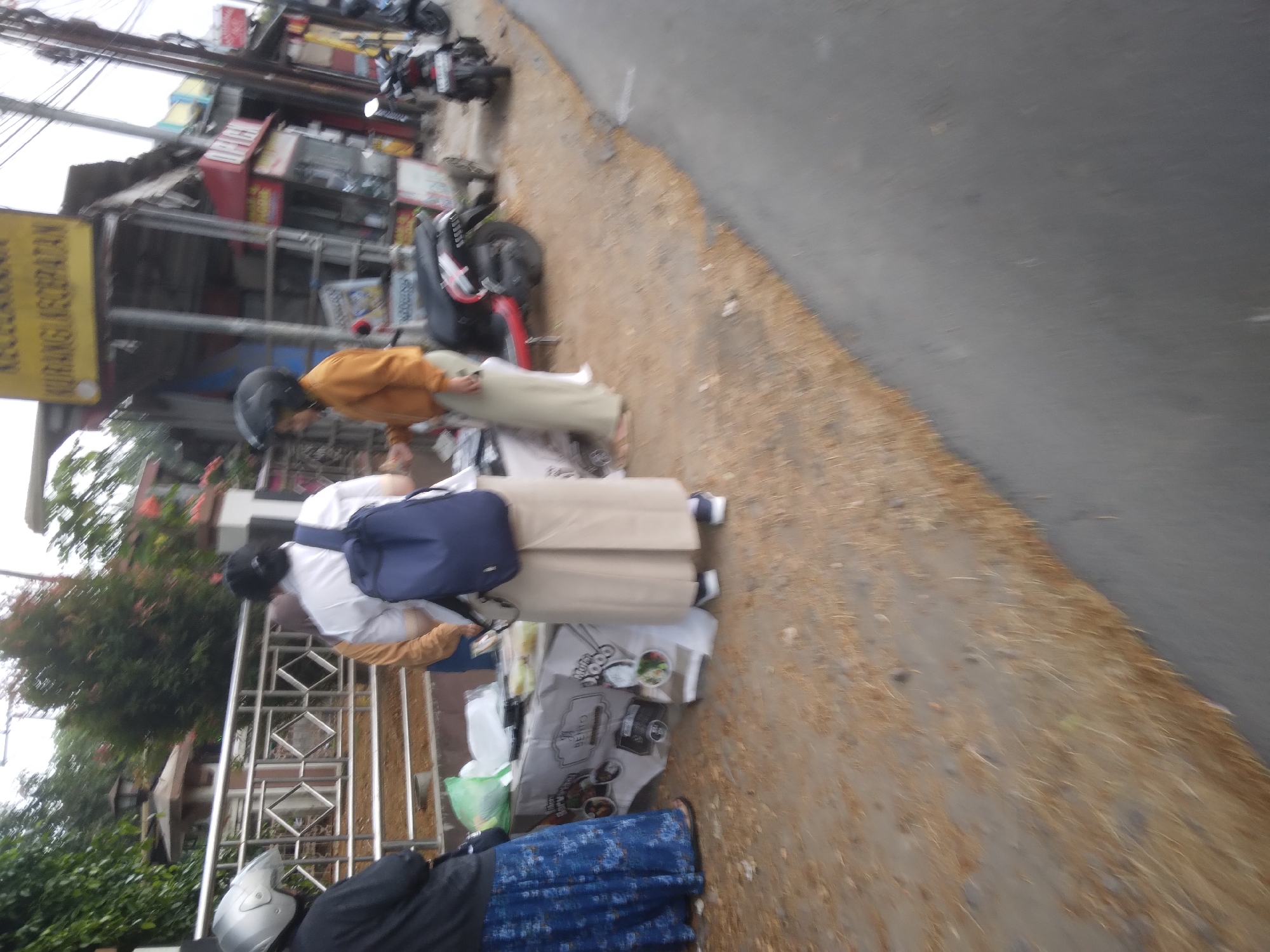 In Desa Pamijen, Kecamatan Sokaraja, Indonesia, a street scene unfolds with a focus on two individuals standing near a food stall. The person on the left, facing away from the camera, wears a black jacket and a blue floral skirt. They are positioned behind a table laden with food containers, some covered with white plastic. A green plastic bag is visible on the table. To the right of this person, a second individual in a white shirt and a long beige skirt faces the food stall, wearing a blue backpack. They appear to be interacting with the stall or its offerings. Further to the right, another person wearing an orange jacket and a light-colored skirt, and a black helmet, stands near a red and black scooter. This scooter is parked behind the food stall. A second, smaller motorcycle is visible further down the street. The ground is covered with dry leaves and dirt, and a paved road runs alongside the scene. Buildings with various signs, including one with yellow lettering reading "KECEPATAN," are present in the background. The overall impression is of a bustling street-side market or food vendor operation.