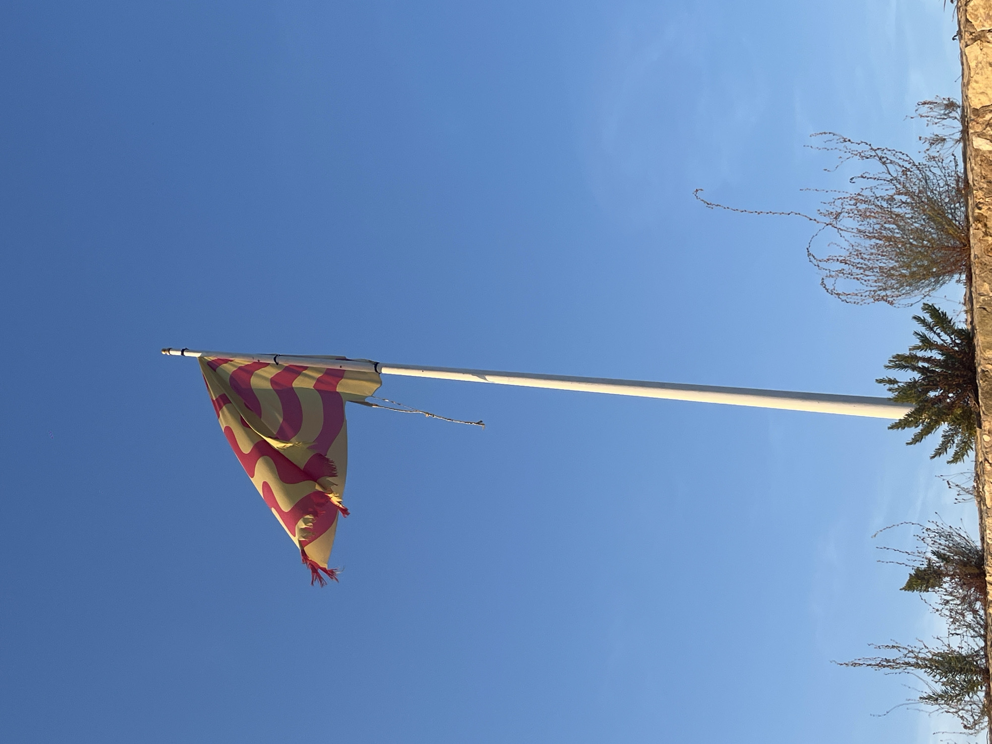 The image shows a flag with pink and yellow stripes fluttering on a pole against a clear blue sky. The flag appears worn, with frayed edges. Below the flag, a low stone wall is visible, topped with sparse green and brown vegetation. A white flagpole stands tall in the center of the frame. The scene is set outdoors, likely in a coastal area given the bright sky and vegetation. This could be a view of the Mediterranean Sea from Tarragona, Spain, a city known for its Roman ruins and seaside location.