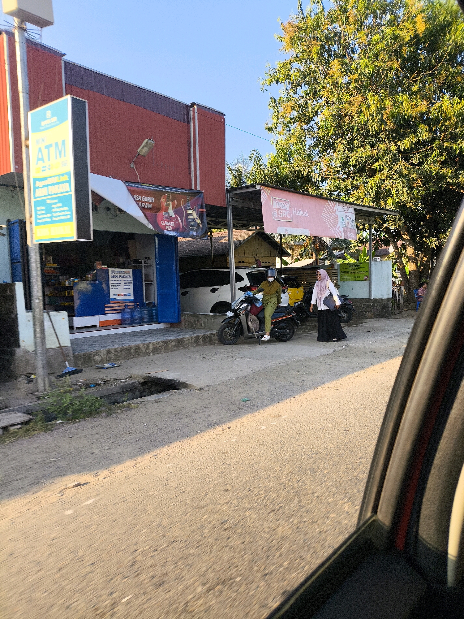 The image captures a street scene in Dulupi, Indonesia, bathed in the warm light of what appears to be late afternoon. In the foreground, the edge of a vehicle's interior is visible, framing the outdoor view. A road stretches out, with a noticeable sunlit patch creating a stark contrast with a shadowed area. To the left, a small shop or building is visible, featuring an ATM sign and various products displayed behind a blue-tinted window. Adjacent to it, a banner hangs, advertising "Toko SRC Haikal."
Two individuals are present near a motorcycle parked on the side of the road. One person, wearing a green outfit and a helmet, is seated on the motorcycle. Another person, dressed in a headscarf and a black skirt and top, is walking away from the motorcycle. A white car is parked behind the motorcycle. Further in the background, a lush green tree with yellow blossoms dominates the upper right portion of the frame. The environment suggests a typical roadside setting in a community.