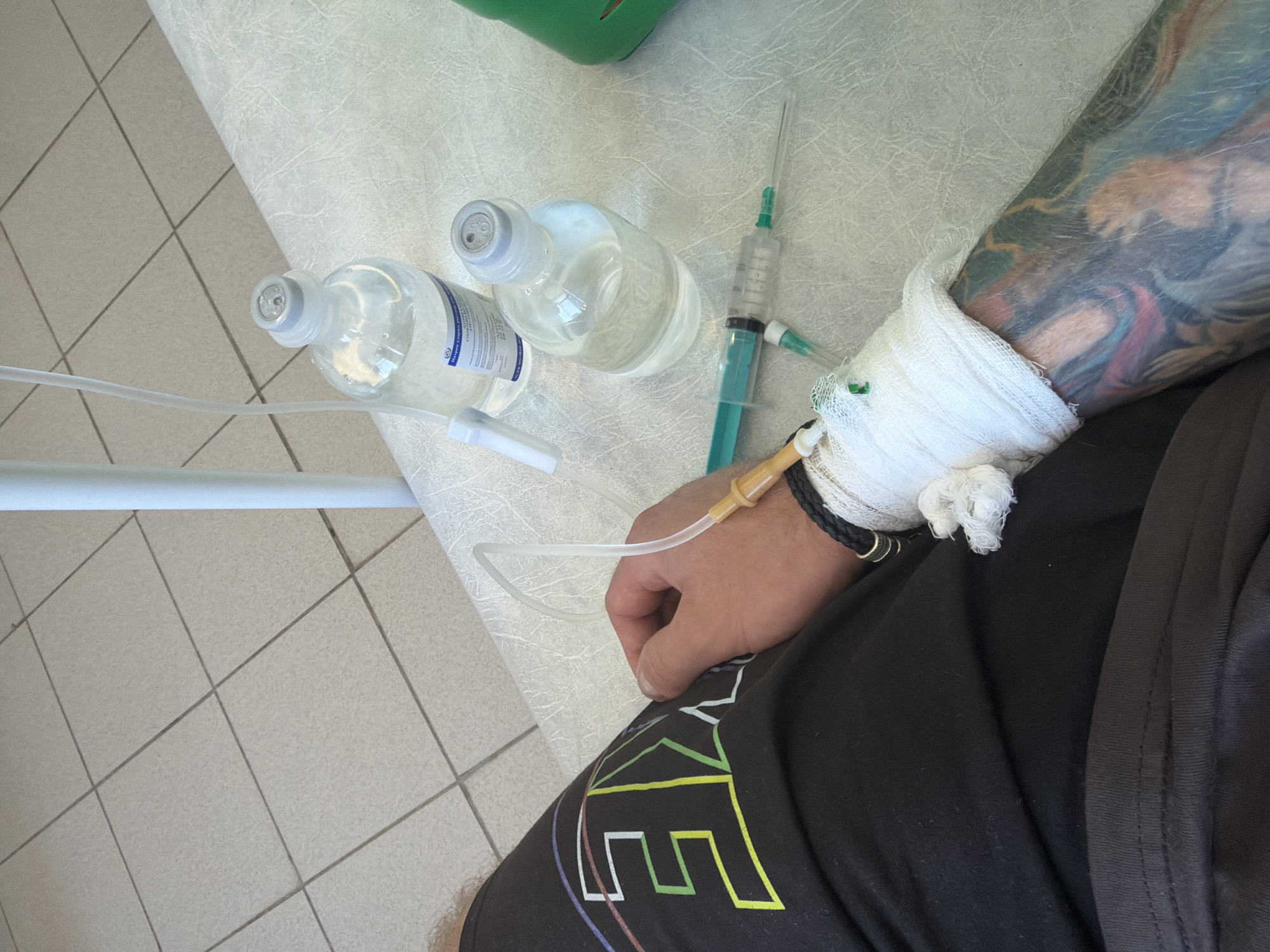 The image depicts a person receiving medical treatment. An IV line is connected to their arm, which is bandaged with white gauze. Two bottles of intravenous fluid are visible on the table beside them. A syringe with green fluid also lies on the table. The person has tattoos on their arm and is wearing a black bracelet. They are wearing black shorts with colorful geometric patterns. The floor is tiled. The location is possibly in a medical facility in the city of Chaikovsky, Russia, based on the information about the IV fluid bottles.