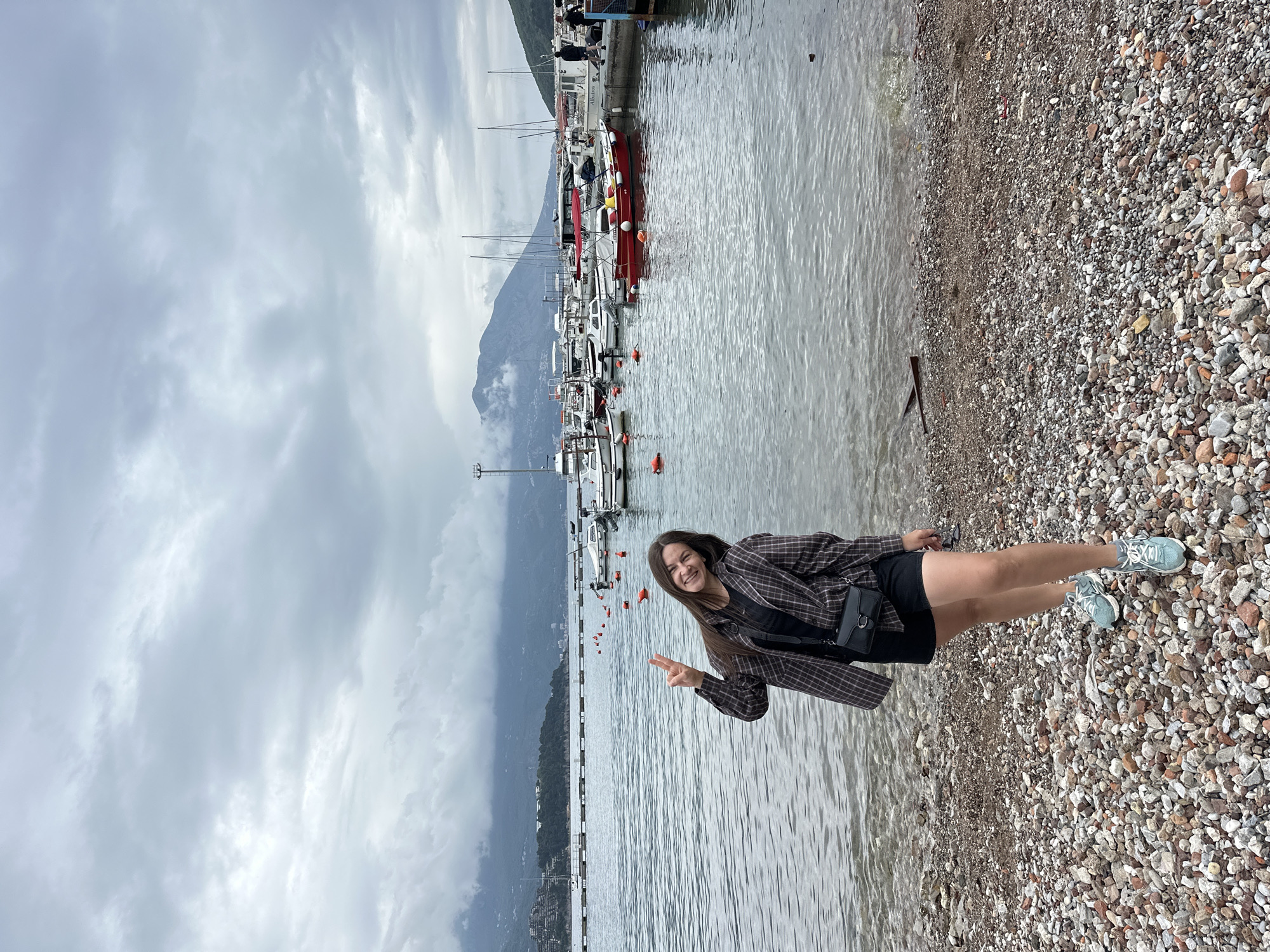 A young woman with long brown hair stands on a pebble beach next to the water, giving a peace sign. She is wearing a black top and shorts with a brown plaid jacket and light blue sneakers. Behind her are docked boats and a mountain range under a cloudy sky. The location is likely near the marina in Budva, Montenegro, given the boats and mountain backdrop.