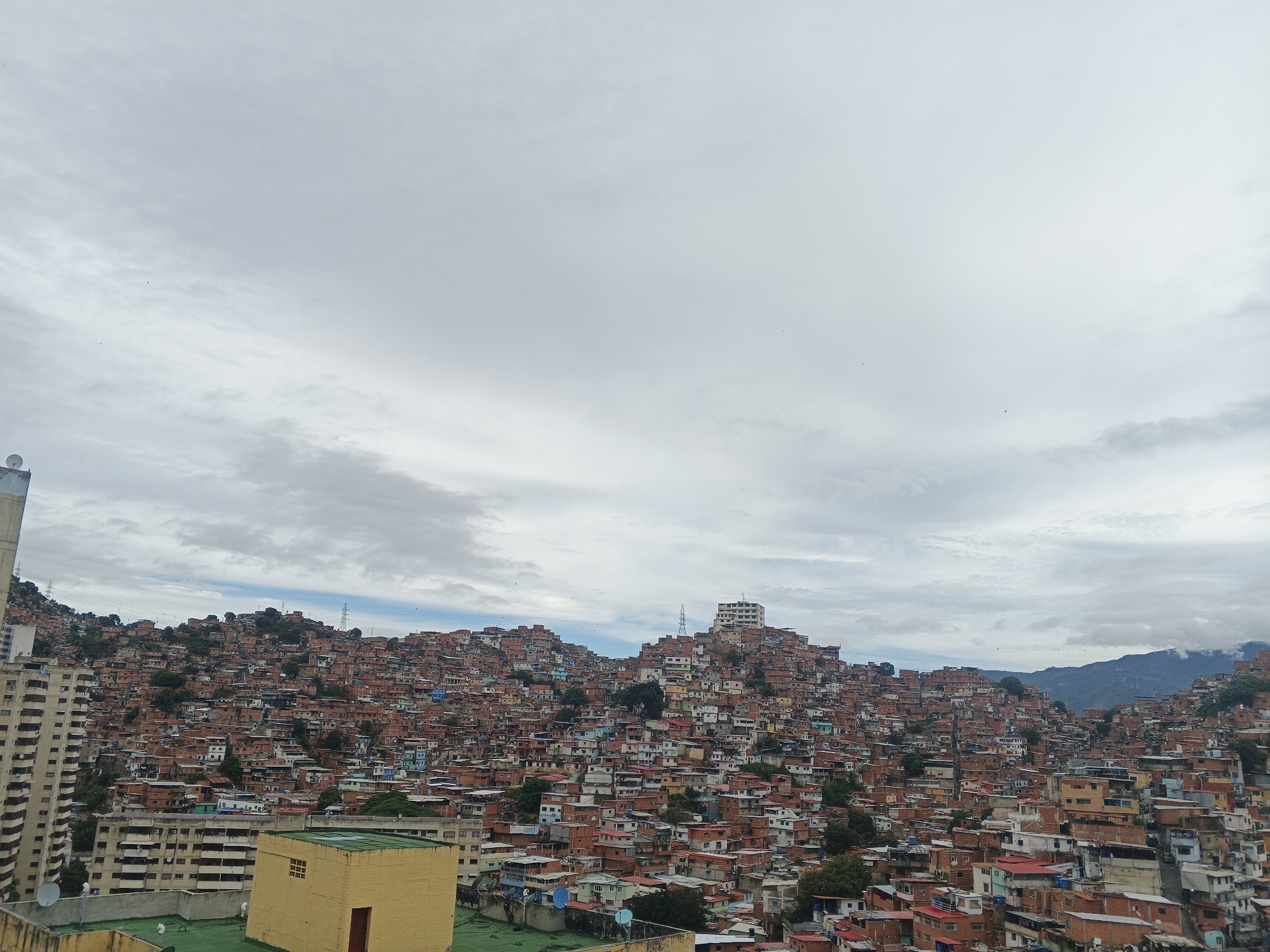 The image depicts a densely populated hillside neighborhood in Caracas, Venezuela, likely a "barrio" or informal settlement. Thousands of small, brightly colored brick and concrete homes are packed together, climbing the steep terrain. The buildings appear to be primarily one or two stories high. In the foreground, a taller building stands to the side, providing a higher vantage point for the photograph. The sky is overcast with thick clouds. The mountains are visible in the distance. The architecture is mostly informal, with a mixture of red brick, concrete, and corrugated iron roofs. There are no people visible in the scene.