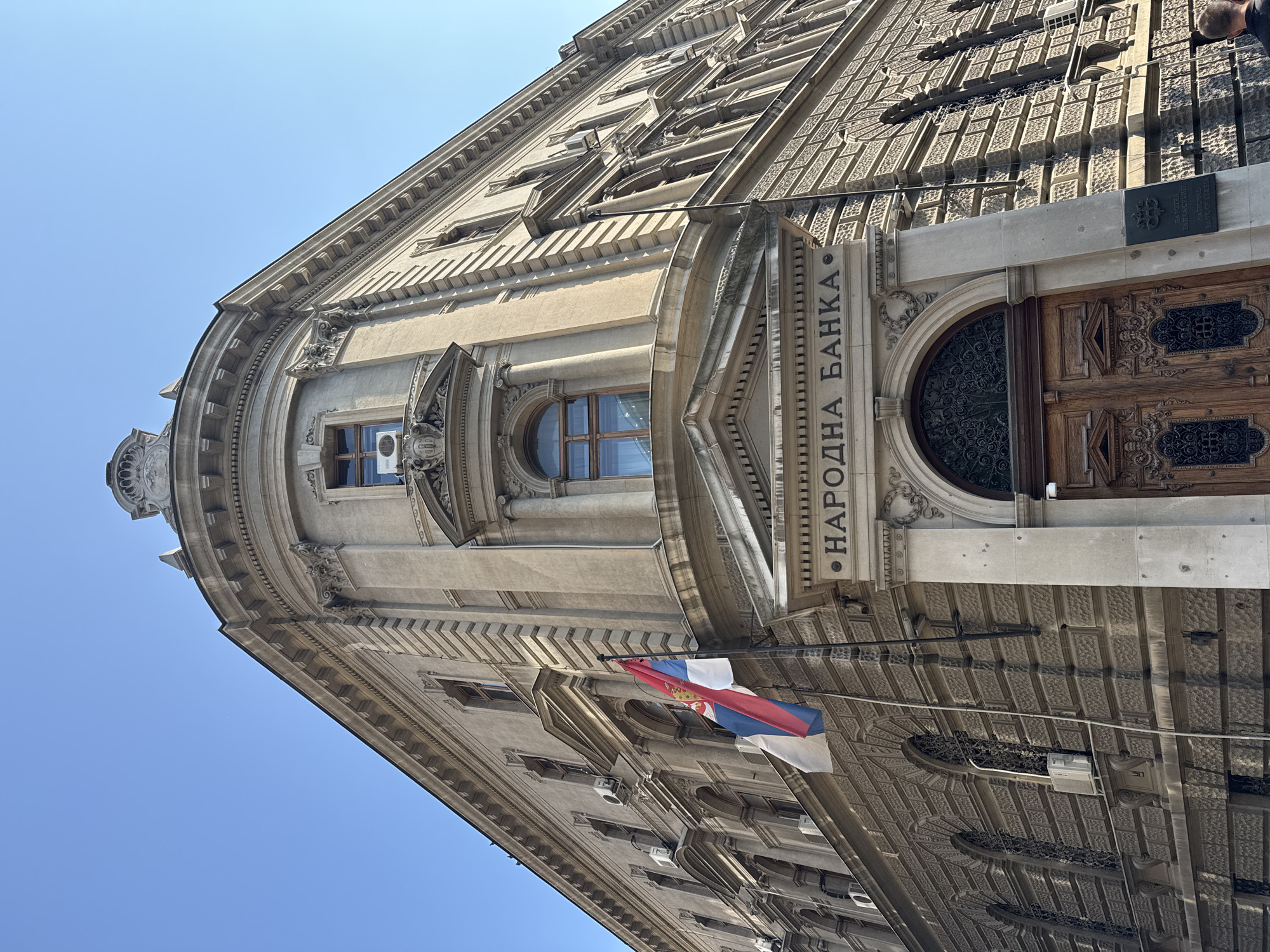 The image shows the exterior of the National Bank of Serbia building in Belgrade, Serbia. The building is a large, ornate structure made of stone, with classical architectural elements such as columns, arched windows, and decorative carvings. A Serbian flag is displayed on a flagpole attached to the building. The sky is clear and blue. The main entrance of the building is visible, featuring a large wooden door with intricate carvings and a decorative metal grille above it. Above the entrance, the words "НАРОДНА БАНКА" are inscribed in Cyrillic. The building's facade is detailed with a variety of textures and patterns, including brickwork and stone cladding. The image is taken from a low angle, looking up at the building. A small portion of a person's head and shoulder is visible in the bottom right corner of the frame.