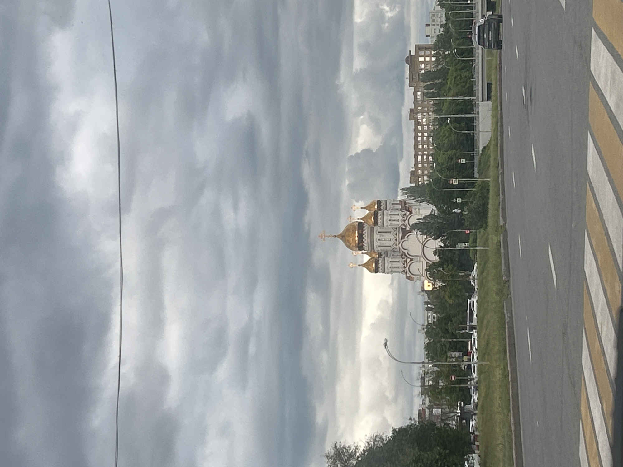 The image captures a view of the Annunciation Cathedral in Приморский, Russia, situated amidst a cityscape. The cathedral's golden domes and white facade stand out against a backdrop of overcast skies with billowing clouds. To the right, a wide asphalt road with pedestrian crosswalk markings dominates the foreground, suggesting an urban thoroughfare. In the distance, buildings and trees line the street, creating a sense of depth within the urban landscape. Streetlights are visible along the road. Cars are seen driving on the road.