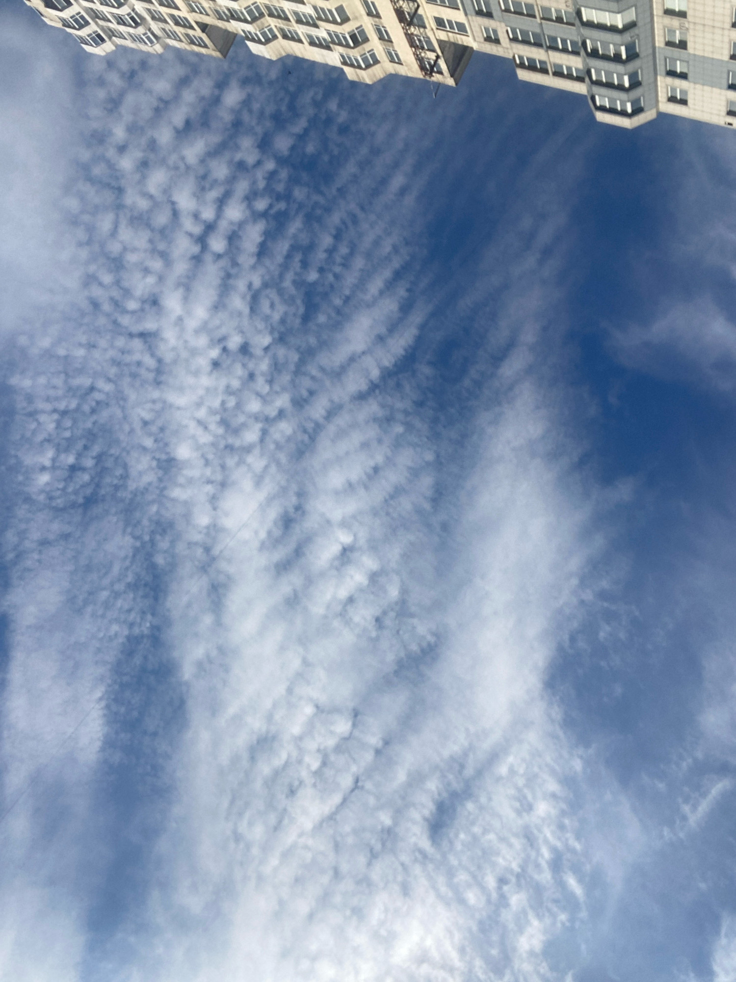 The image depicts a view looking upwards towards the sky and the upper portion of a multi-story building. The sky is a deep blue and is filled with wispy white clouds. The building appears to be modern, with rows of windows and a light-colored facade. The orientation of the image suggests it was taken from a ground-level perspective looking up, or from a lower floor of another building. The location is identified as Chornomorsk, Ukraine. There are no people or discernible actions visible in the image.
