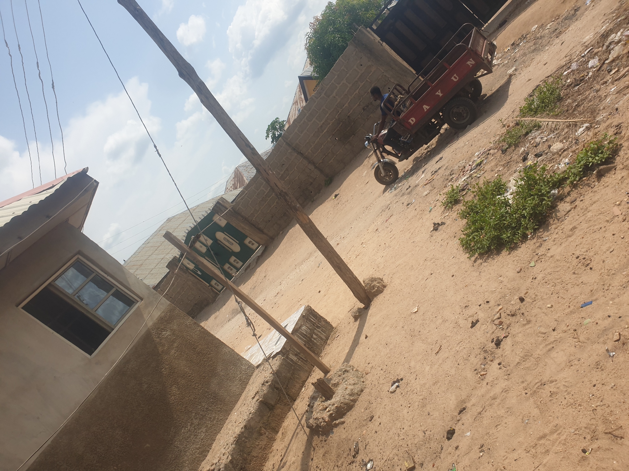 The image shows a dirt road in an urban area. There is a three-wheeled vehicle with the name "DAYUN" on its side being driven by a person. A building with a window is on the left, and a concrete wall is on the right. A wooden pole stands in the foreground. The sky is visible with some clouds.