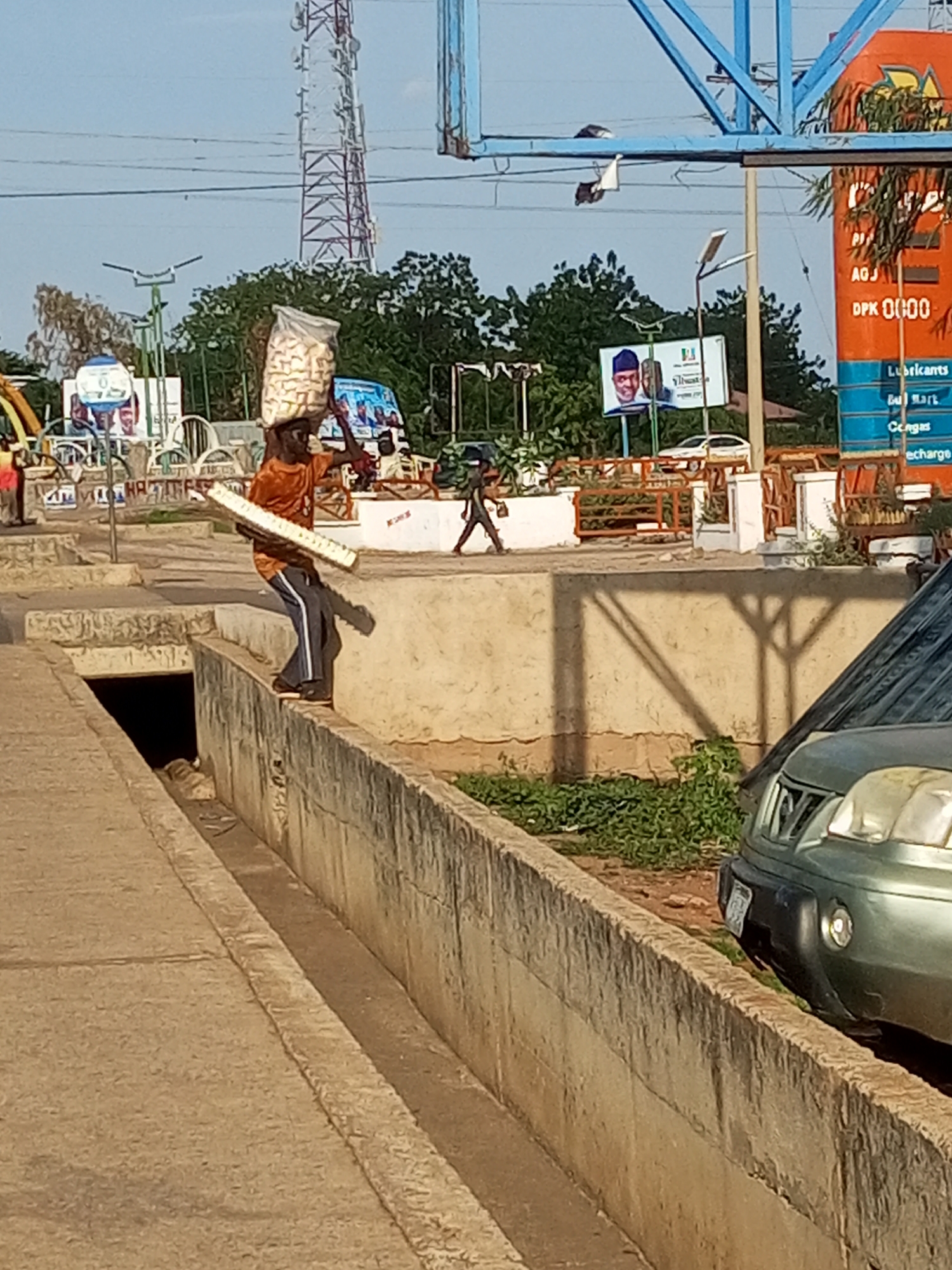 The image depicts a scene in Gombe, Nigeria, under a clear, bright sky, suggesting daytime. The primary subject is a young man carrying a large, clear plastic bag filled with what appears to be snacks, balanced on his head. He is walking along the top of a concrete structure, possibly a wall or the edge of a drainage ditch, with his body leaning slightly backward to maintain his balance. He is wearing an orange t-shirt and dark pants with a white stripe. He is also carrying a long, cylindrical object, possibly a rolled-up mat or poster, in his right arm.
In the background, several elements provide context. A tall telecommunications tower stands prominently, a common sight in urban and semi-urban areas. Several trees with lush green foliage are visible, suggesting a warm climate. There are also signs with text, including one that reads "AGJ DPK 0000". Another sign features images of two men in traditional attire, possibly political figures, and text in what appears to be Hausa and English. Further in the background, a green SUV is parked, and a light blue metal structure, possibly part of a playground or an advertisement display, is visible. The overall setting appears to be a busy public area, perhaps near a market or a transit hub, with other individuals and structures hinting at daily life. The activity appears to be the man's daily work of transporting goods.