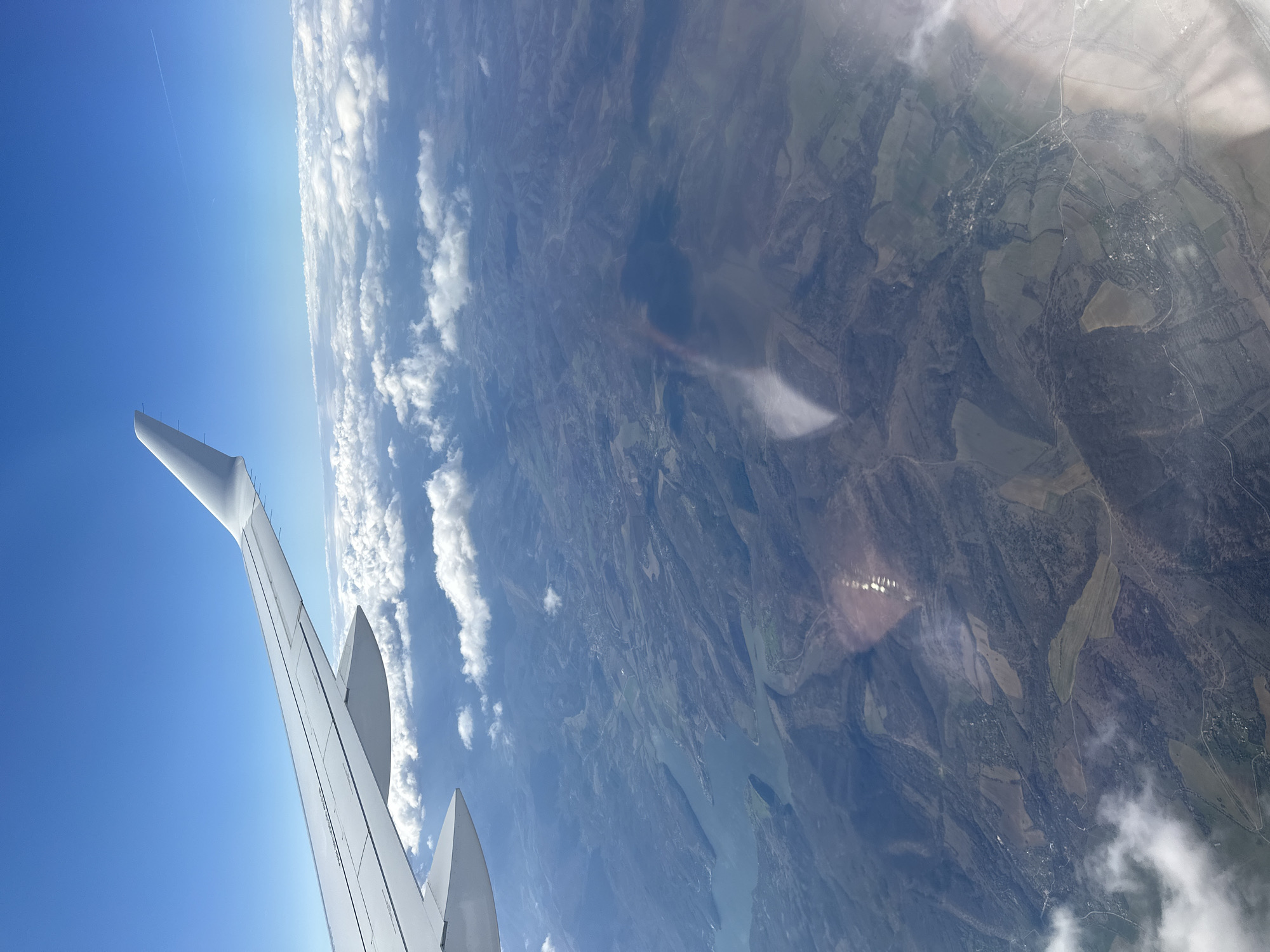 The image shows a view from an airplane window, looking down at the landscape. In the foreground, a portion of the airplane's wing is visible against a clear blue sky and a bank of clouds. Below the clouds, a mosaic of fields, forests, and possibly a river or lake can be seen. The overall tone suggests a rural area, possibly near Kosharite, Bulgaria, though the exact location cannot be determined with certainty. There are no people visible in the image.