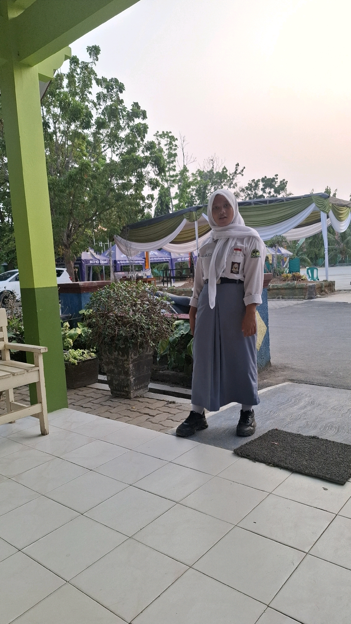 A young person wearing a white hijab and a school uniform stands on a tiled patio. The uniform consists of a white long-sleeved shirt with patches on the chest and a gray skirt. They are wearing black sneakers. In the background, there are trees, a tent with decorative draping, and a white car parked. The location is the grounds of a school in Bandar Lampung, Indonesia, likely during an outdoor event or assembly. The sky is overcast with a hint of sunset.
