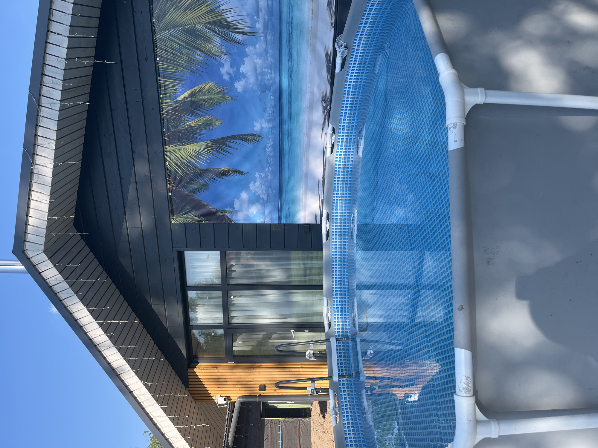 The image shows a portion of a backyard in Новая Чернушка, Russia. A prefabricated above-ground swimming pool with a blue patterned liner and grey frame is partially visible. Adjacent to the pool is a modern-style house with a section featuring black wood siding, a large window, and string lights along the roofline. A portion of the house has a wall with lighter, horizontally placed wood siding. A backdrop depicting a beach scene with palm trees and blue sky is positioned on the house, serving as a poolside decoration. Dirt and what appears to be landscaping materials are visible near the base of the house. The sky is clear and blue.