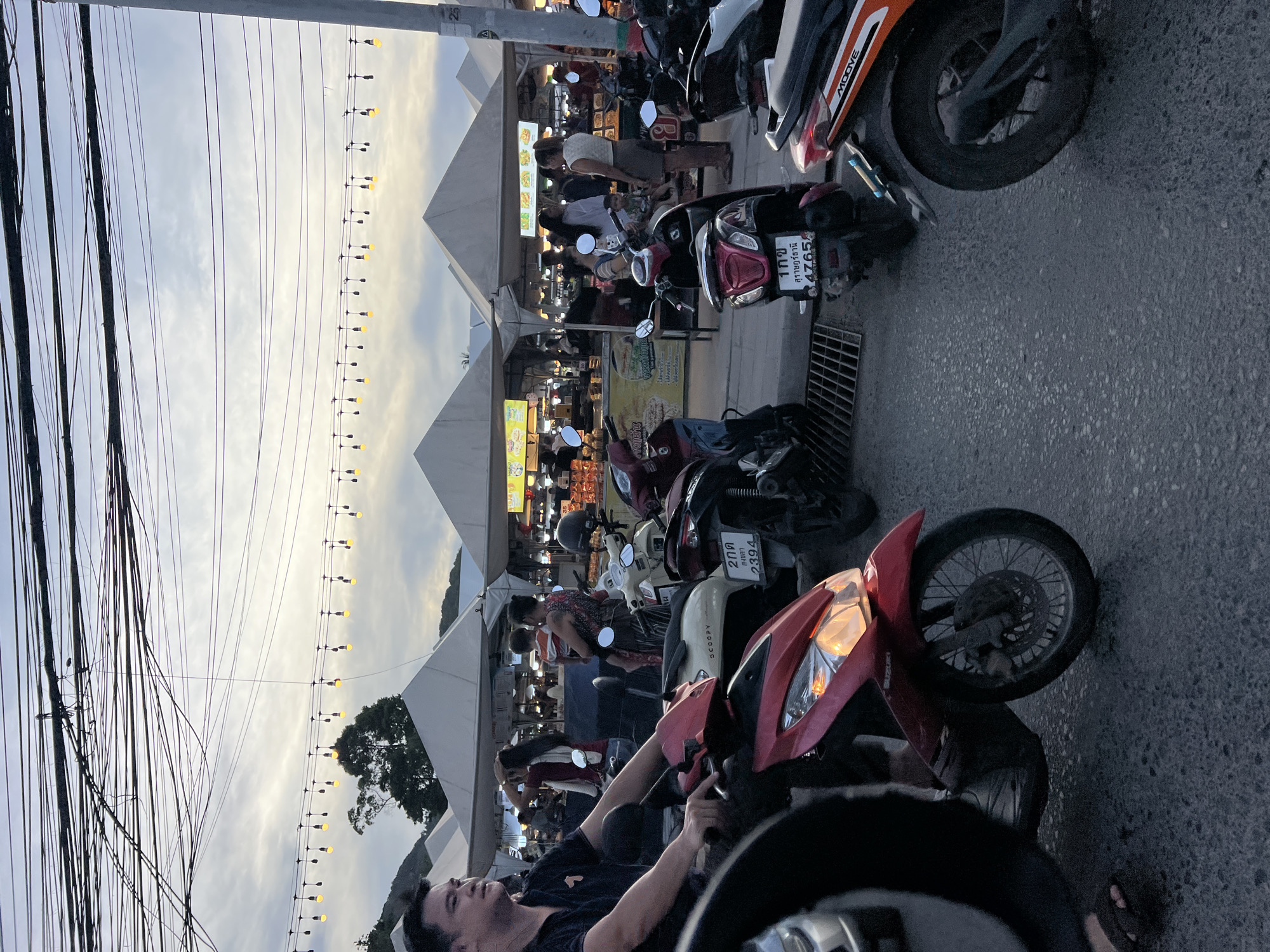 The image shows a street scene in บ้านบางรักษ์, Thailand. Numerous parked motorcycles line the right side of the frame, partially obscuring food stalls beneath white triangular tents. People are gathered at the food stalls. Overhead, strings of lights extend across the street. A man is seated on a motorcycle, his arms resting on the handlebars. A web of power lines runs along the left side of the frame.