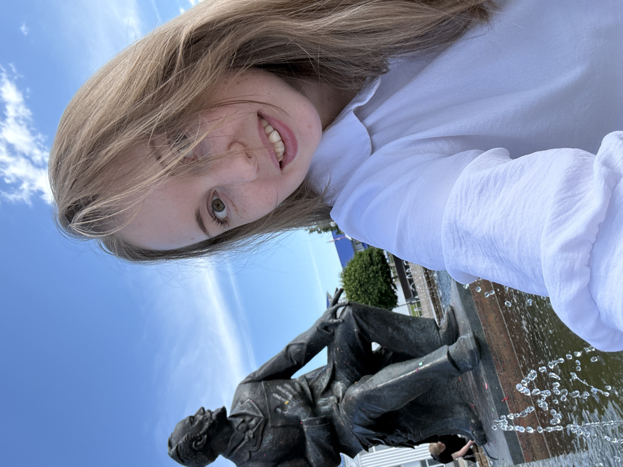 A young woman with light brown hair and light eyes is captured in a close-up selfie, smiling subtly towards the camera, with her hair partially obscuring her face. She is wearing a white collared shirt. Behind her, slightly to the left, is a bronze statue of a seated, bearded man in a suit, holding an object to his chest. The statue shows visible colorful specks. It is positioned on a dark stone base, which is part of a fountain with water actively spraying upwards, creating visible droplets in the foreground. In the mid-ground, a person in a dark shirt is partially visible crouching near the base of the statue. The background features a bright blue sky with sparse white clouds, green foliage, a white building with vertical panels, and a blue-roofed structure. The scene is outdoors in the city of Unknown, Unknown.