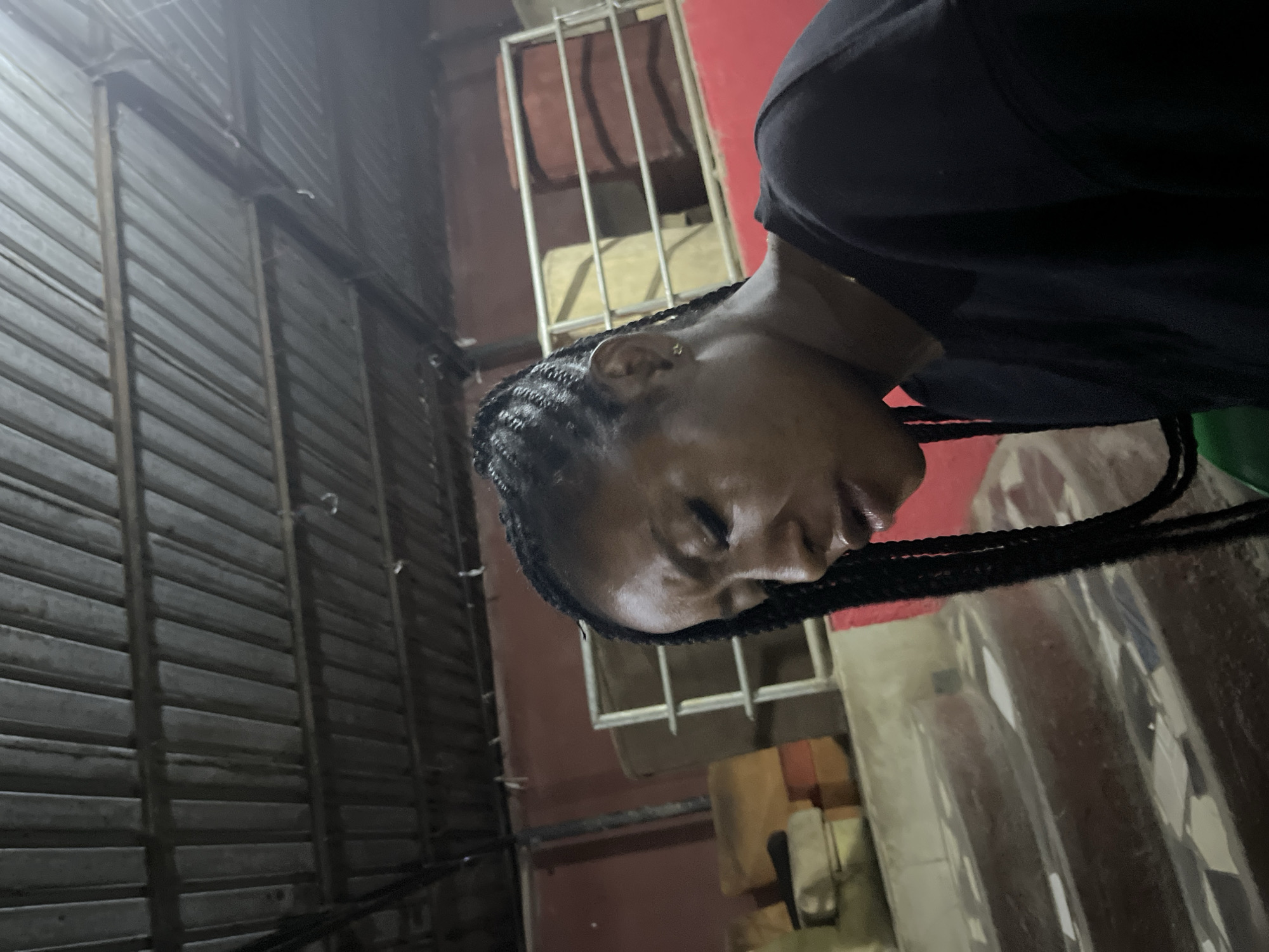 A medium shot shows a young Black woman in the foreground, her head bowed and eyes closed, with her face angled downwards to the right. She has dark, braided hair that cascades down her left side. She is wearing a black top. The background is dimly lit and appears to be an outdoor or semi-enclosed space with a corrugated metal roof overhead. To her right and behind her, there are some stacked, cushioned objects in tan and orange colors, and a red wall. Further back, there is a metal railing structure with what appear to be cylindrical objects behind it, one in a reddish-brown color. In the lower left quadrant of the image, there are tiered steps with a mosaic-like pattern on them, and the edge of a green object is visible. The scene suggests a casual, possibly rustic setting.