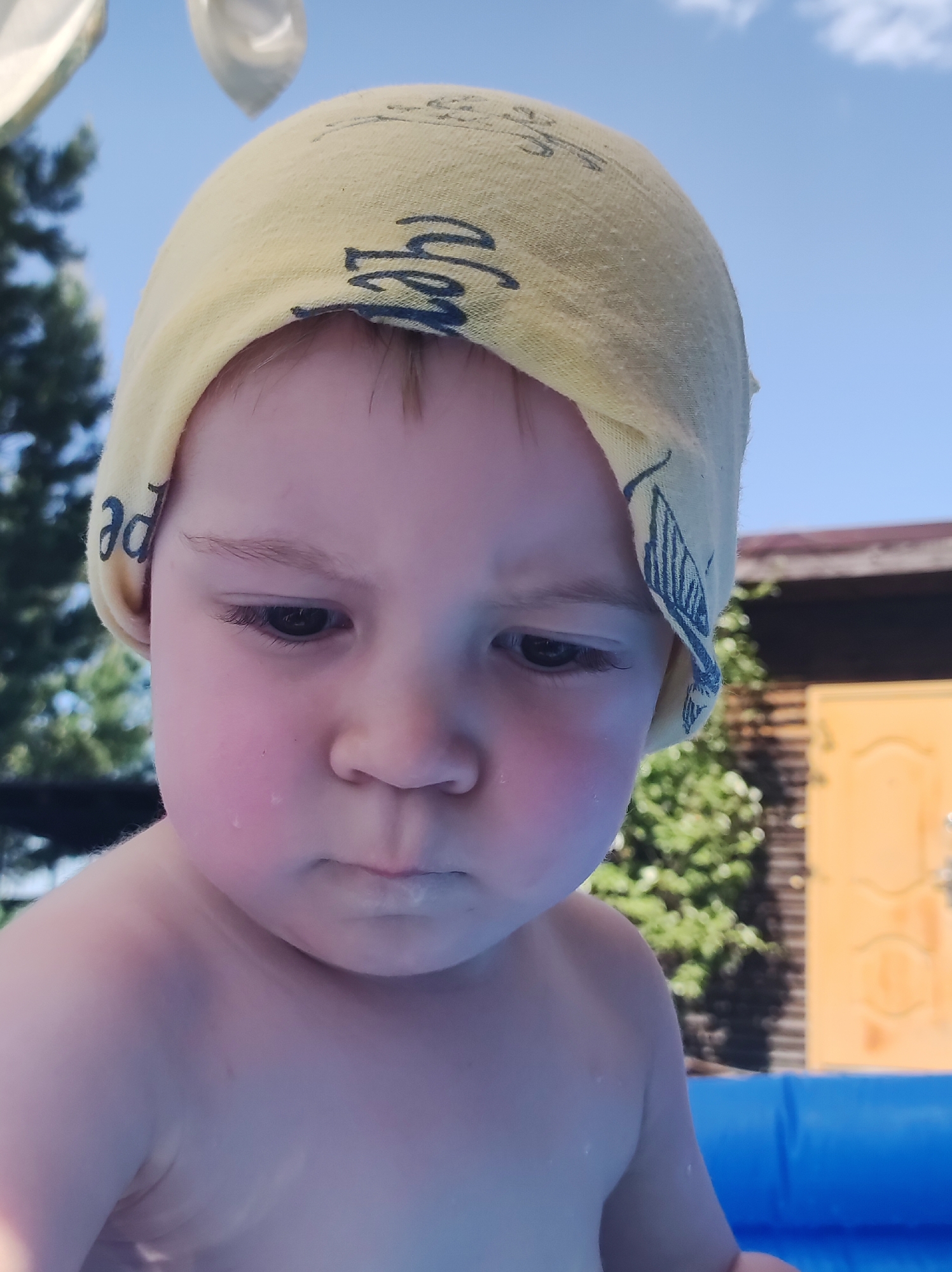 A young child with fair skin and light-brown hair is looking down. The child is wearing a yellow head covering with a blue pattern. The child's face has flushed cheeks and appears to be wet. The child's torso is visible. In the background, there are trees, a blue sky with clouds, and a light-brown building. There is a partially visible blue inflatable pool.
I am not able to find information that would confirm the location is Фоки, Russia.