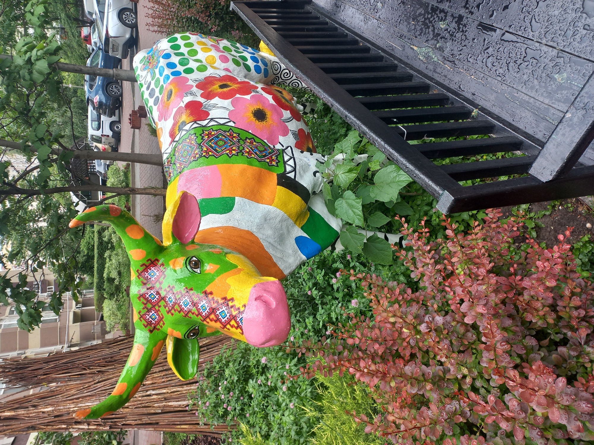 A vibrantly painted cow sculpture stands in a flower bed next to a black staircase. The sculpture is adorned with colorful polka dots, floral patterns, and geometric designs. It has a pink nose, green and orange horns, and green skin with orange spots. In the background, parked cars line a street beside trees. The scene is located in the city of Kyiv, Ukraine.