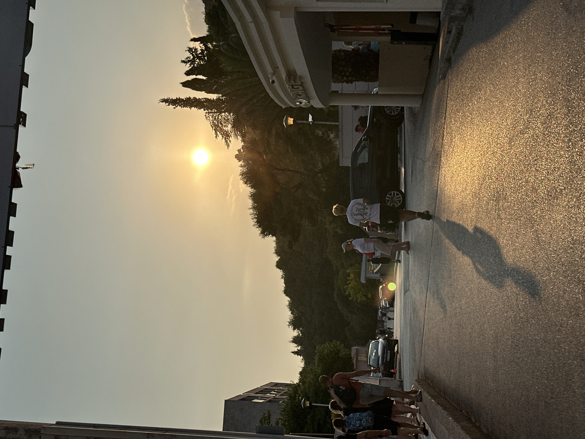 The image depicts a street scene in Bečići, Montenegro, at sunset. The sun is low on the horizon, casting a warm golden light on the wet asphalt road and creating long shadows of people.
In the foreground, several individuals are walking along the left side of the road. One person, facing away from the camera, is wearing a white t-shirt with graphics and dark shorts, walking towards the right. Their shadow is prominently cast on the road. To the left of this individual, a group of people are standing or walking, including a person with a backpack.
The road itself appears to be a two-lane street, with cars parked or driving in the distance. A dark-colored SUV is visible in the middle ground, and other vehicles are further down the street.
On the right side of the image, there is a building with a curved facade. A sign above the entrance reads "GARAGE". A CCTV camera is mounted on the building.
The background features a lush green hillside covered in trees, with a few tall cypress trees rising above the canopy. The sky is a gradient of pale yellow and blue, typical of a sunset. The overall atmosphere is peaceful and warm, with a sense of people enjoying the evening.