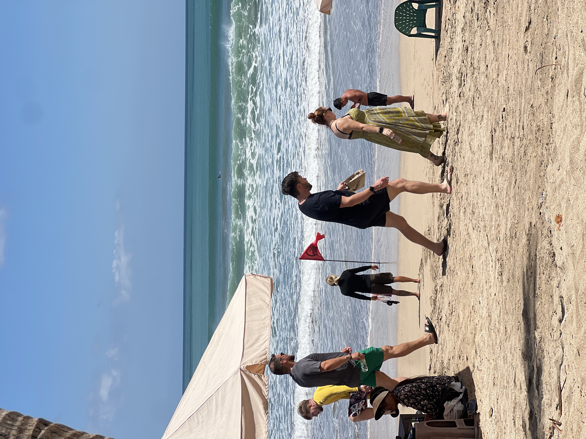 The image depicts a sunny day on Kuta Beach, Indonesia, with several people enjoying the sand and surf. In the foreground, a woman in a long, green and yellow patterned dress walks on the sand, holding a water bottle. To her left, a man in black shorts and a t-shirt walks with a bag. Further left, a man in green shorts and a dark grey t-shirt walks, holding a bottle of water. Behind him, a man in a yellow long-sleeved shirt walks with a patterned pair of shorts. To the right, a person in black shorts and a black shirt stands facing away from the camera. In the middle ground, a person in black stands near a red flag planted in the sand. The ocean features gentle waves, and a distant surfer can be seen on a board. A large, cream-colored umbrella provides shade in the left foreground.