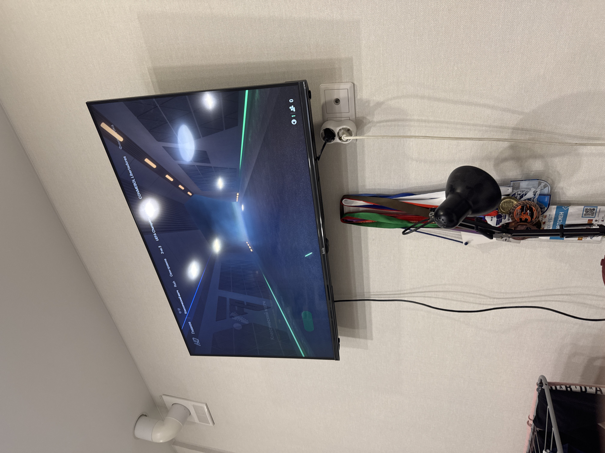 The image captures an indoor scene, likely in a room in Kazan, Russia, focusing on a wall-mounted flat-screen television and its immediate surroundings. The wall is covered in light-colored, textured wallpaper.
The central subject is a large, black-framed TV displaying a dark interface with bright, stylized lights and green/blue lines, suggesting a video game or sports simulation, possibly football. The screen shows menu options in both English and Russian. Visible text includes "0 1 0", "Главное" (Main), "Классический матч" (Classic match), "Rush", "Свои правила" (Your rules), "3 на 3", "UEFA Champions", "CONMEBOL Libertadores", "Фина" (Final), "tuvio", and "HD".
To the right of the TV, a white electrical outlet cluster is plugged into the wall, powering the television and another device. Above it is another single white outlet. Below these outlets, a black table lamp with a dome-shaped head rests on a shelf. This shelf is cluttered with various items, including multiple medals with red, green, and blue ribbons, small circular objects (possibly coins or badges), and some printed materials. Text visible on these items includes "CK-PECT" and "IPTCHEN". A black cable runs horizontally across the wallpaper above the shelf.
In the bottom left of the frame, a white vent or duct system is visible. The bottom right shows a sliver of metal-framed furniture. The photo is taken from an elevated, angled perspective.