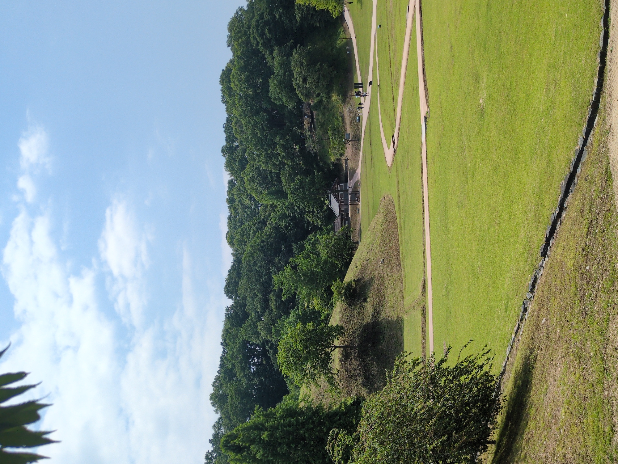 The image shows a landscape in Gongju-si, South Korea. The scene includes a clear blue sky with scattered white clouds. Lush green trees line the perimeter, giving way to an expansive grassy area intersected by winding dirt paths. Several people can be seen walking along these paths. There is a small building with a traditional Korean roof visible in the distance. A small mound covered in grass is positioned near the edge of the trees. A stone barrier separates the grassy area from another grassy area.