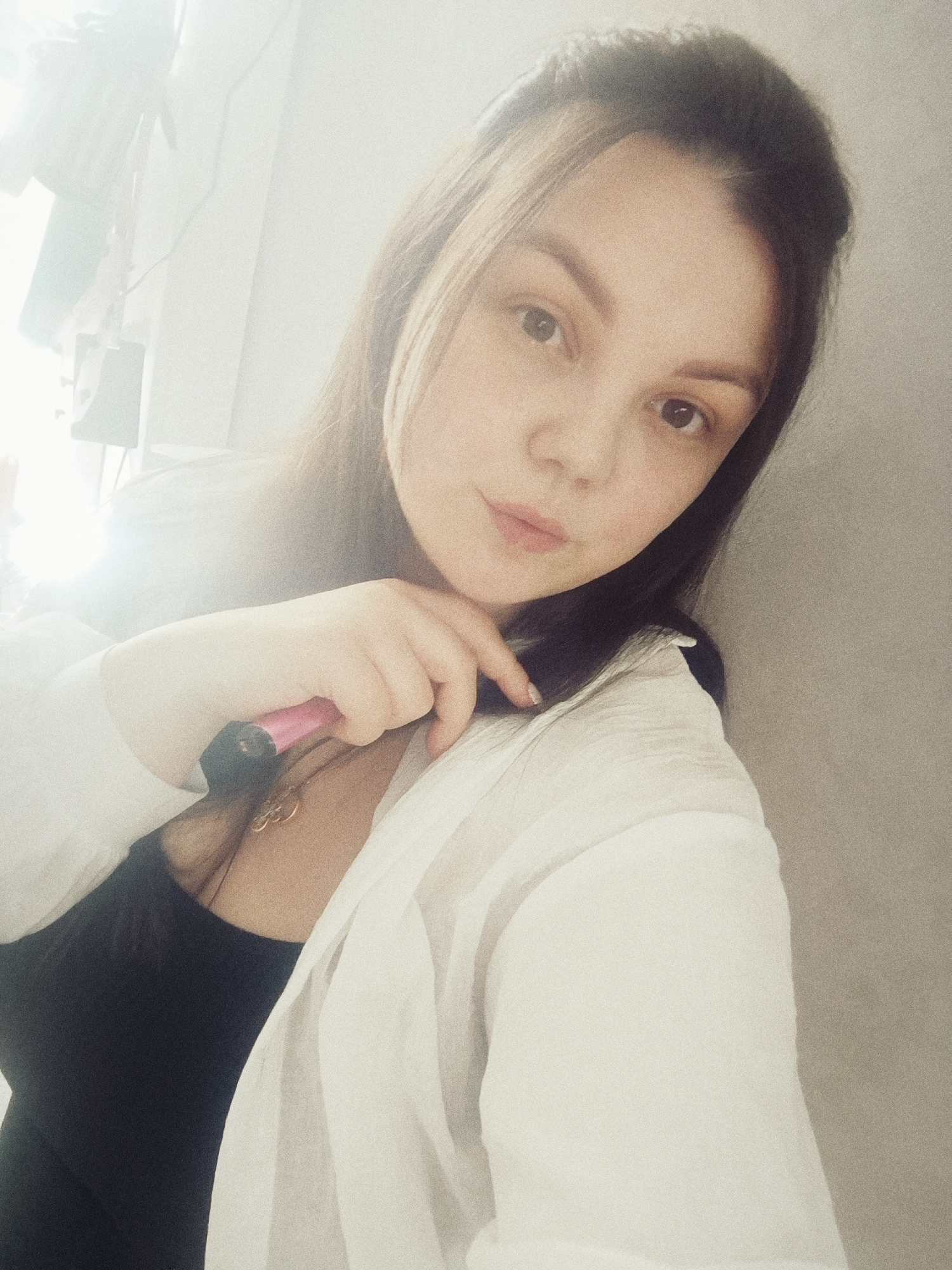 A woman is posing for a selfie indoors. She is fair-skinned with dark hair, wearing a black top under an unbuttoned white shirt. She holds a pink vape pen near her neck. A necklace is visible under her shirt. The background appears to be a wall and a blurry section possibly containing light-colored furniture or fixtures. The photo has a slightly grainy quality. The location, Chaikovsky, Russia, cannot be determined from the visual information in the image.