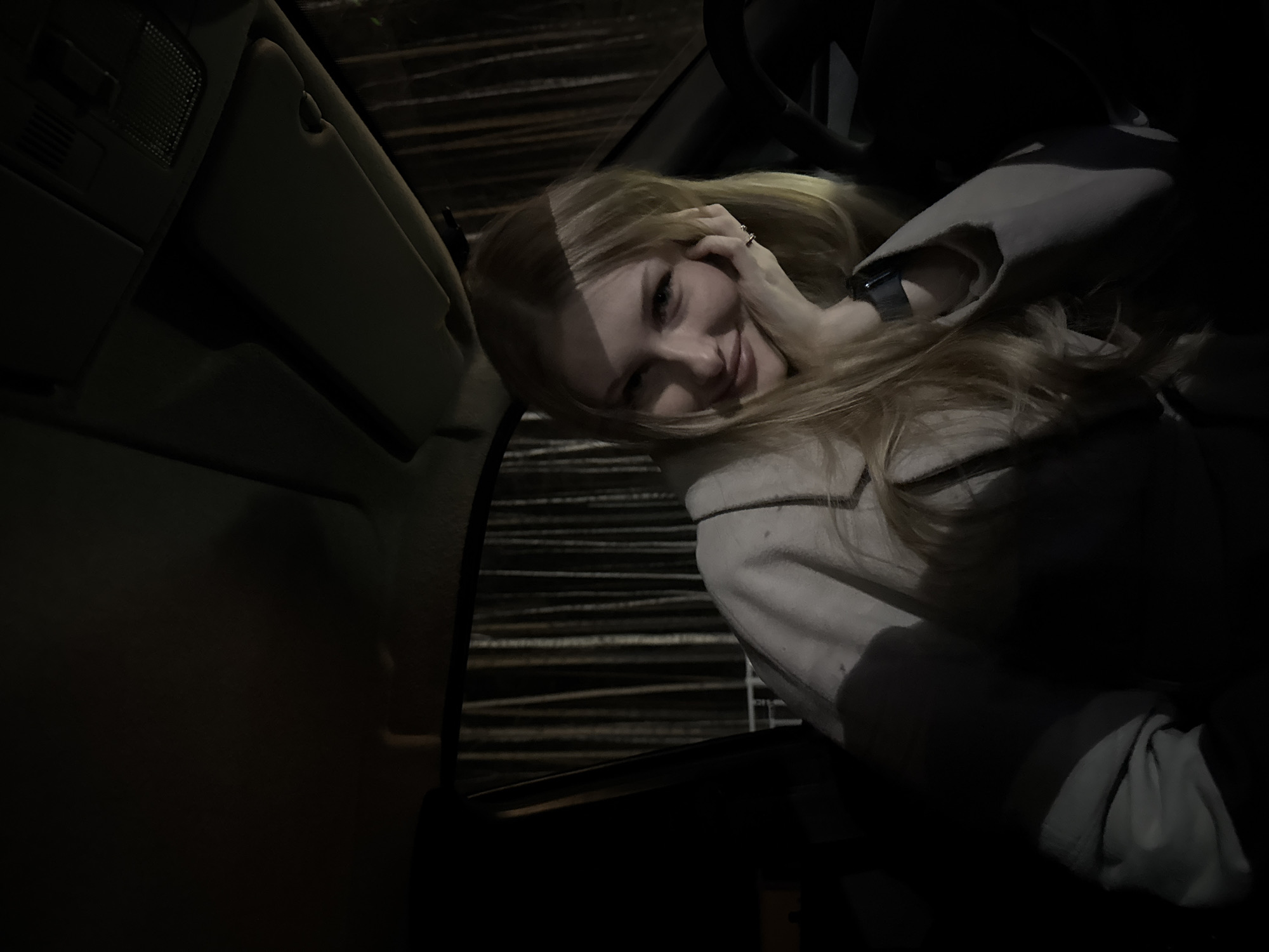 This image captures a young woman inside a car at night in Perm, Russia. She has long, flowing blonde hair, partially illuminated by a low-angle light source, casting a stripe of light across her forehead and cheek. She is looking towards the camera with a playful expression, featuring a slight smile or smirk and what appears to be a wink. Her left hand is raised near her face, with a ring visible on one finger, while a dark smart watch is on her left wrist. She is wearing a light-colored, possibly off-white or light grey, coat or jacket.
The setting is a car interior, with parts of the door panel, roof console, and a sun visor visible in the dimly lit foreground. The background, seen through the car window, is dark with horizontal light lines, suggesting artificial lighting on a distant building facade or industrial structure at night. The lighting is very dim, creating strong contrasts and deep shadows throughout the scene, especially on the car's interior elements. The image is tilted, adding a dynamic composition. There is no visible text.