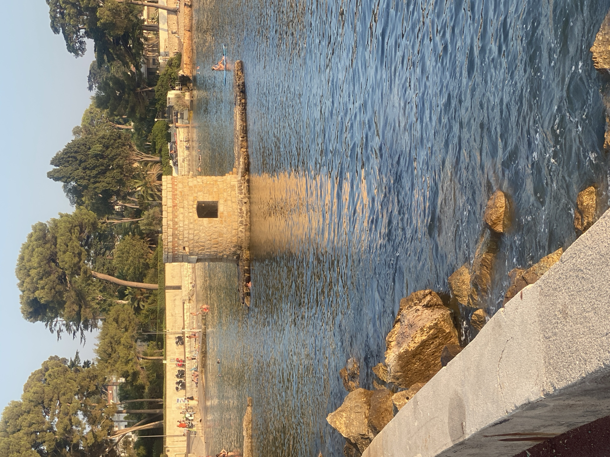The image depicts a serene coastal scene in Antibes, France, likely during late afternoon given the warm, golden light illuminating the stone tower and the water. In the foreground, a concrete wall and large, sun-drenched rocks line the edge of the clear blue water. The water itself is calm, with gentle ripples and the reflection of the stone tower, which stands on a small rocky outcrop in the middle ground. This tower, constructed of rough-hewn stone, has a single, dark rectangular window.
Further back, a sandy beach is visible with numerous people enjoying the water and the shore. Some are sunbathing, while others are wading or swimming. A few individuals are seen standing or sitting on the beach, with some motorcycles parked behind them. In the distance, lush green trees and vegetation line the coastline, interspersed with buildings and homes. A person is visible on a paddleboard in the water on the right side of the frame. The sky is a clear, pale blue. The overall impression is one of a tranquil, sun-drenched Mediterranean afternoon.