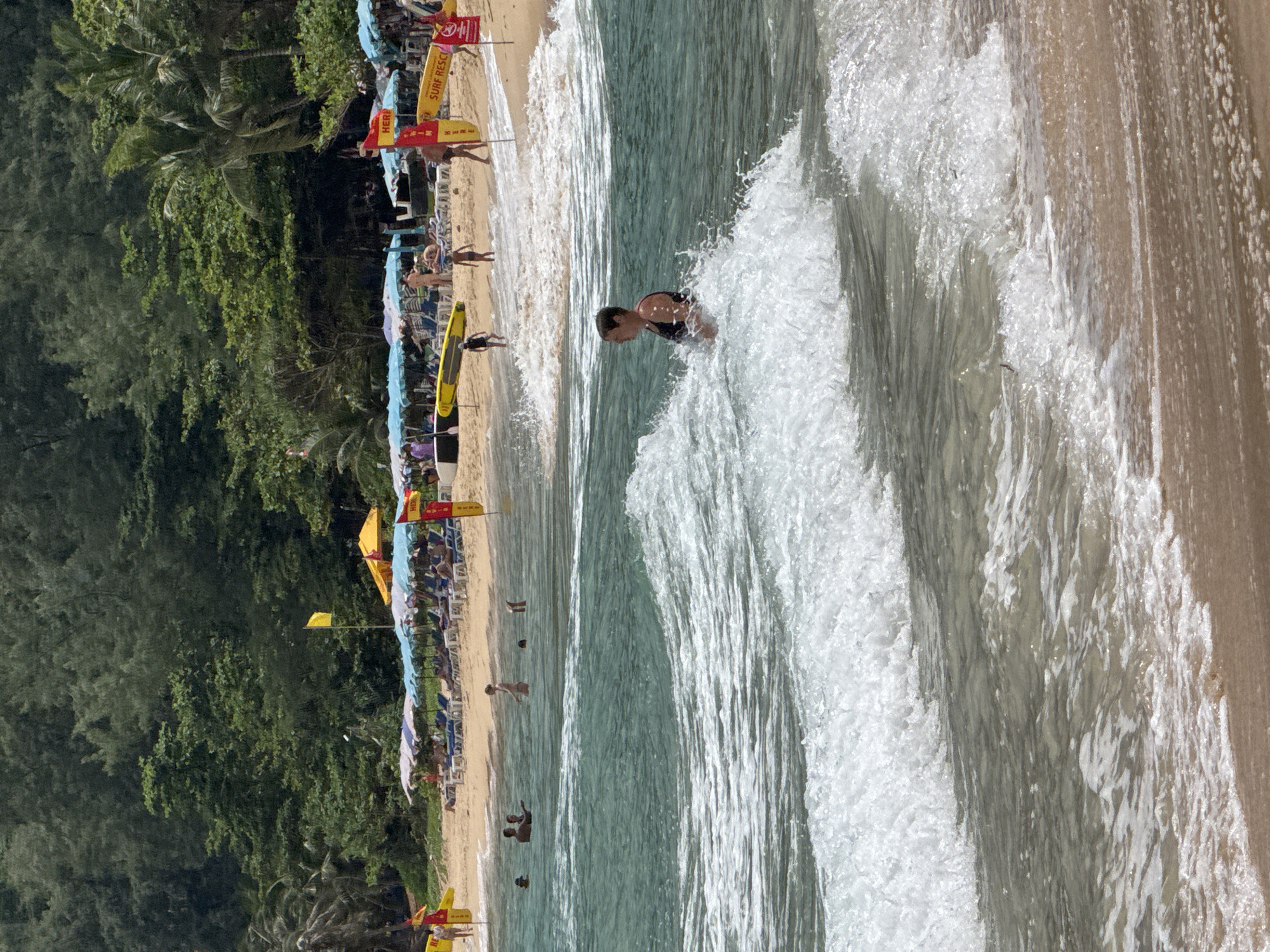 An overhead shot captures a beach scene in an unknown location. The foreground is dominated by a foamy wave breaking on a sandy shore. In the middle ground, a woman in a dark swimsuit stands in the shallow water, facing away from the camera. Further back, several people are wading in the ocean. The background reveals a crowded beach with numerous umbrellas and lounge chairs. Lifeguard flags and surfboards are visible on the sand, suggesting a designated swimming area. Lush green trees and palm trees line the edge of the beach, forming a dense backdrop. The sky is a clear blue.