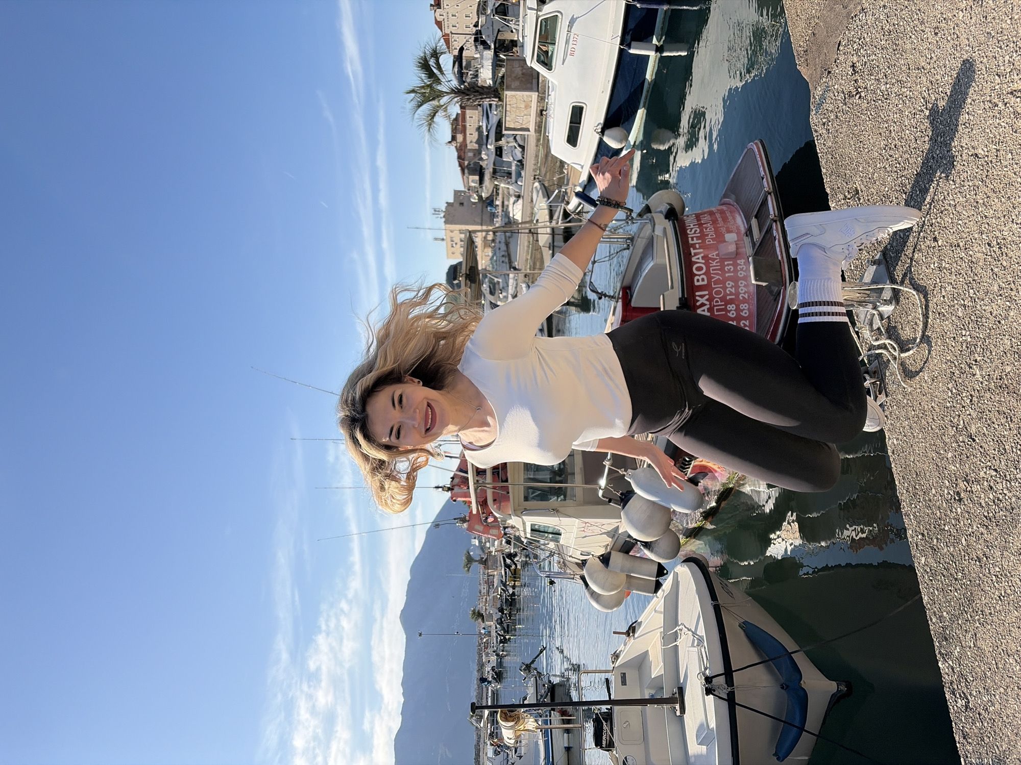 The image shows a woman standing on a dock in Budva, Montenegro. She is wearing a white long-sleeved shirt and black leggings. She is wearing white sneakers and socks with stripes. She has blonde hair. She is smiling and posing for the camera. There are boats docked in the background and a mountain in the distance. The OCR indicates that the boats offer taxi services for boat trips and fishing. The boats are in the harbor of Budva, Montenegro. The sky is blue. The photo appears to have been taken on a sunny day. The woman is standing on a concrete dock.