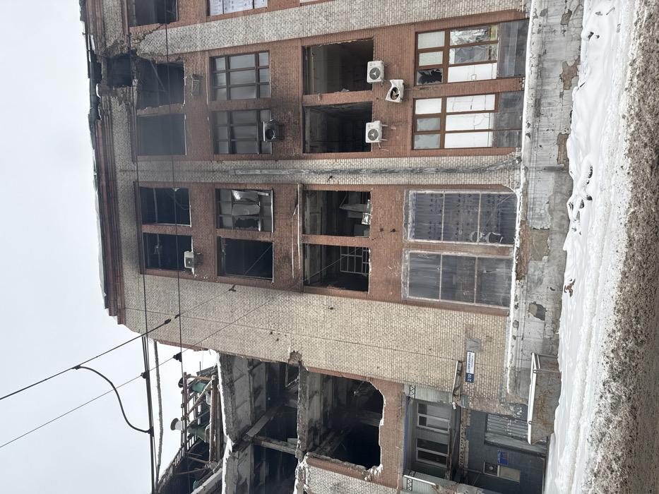 The image captures a severely damaged multi-story building under an overcast sky, suggesting a winter setting due to visible snow and ice. The building appears to be of an older construction, featuring a facade made of reddish-brown and light beige or cream-colored bricks.
Significant structural damage is evident across multiple sections of the building. On the left side of the upper floors, a large portion of the structure appears to have collapsed or been blown out, leaving gaping holes where walls and ceilings once were. Many windows throughout the building are shattered, broken, or completely missing, revealing the dark interiors of the rooms. Some window frames are bent and twisted, and there are areas of dark discoloration around certain openings, possibly indicating charring from a fire. Several external air conditioning units are still attached to the facade, some hanging precariously or appearing damaged.
In the foreground, utility lines or power cables stretch across the frame. To the right of the building, a vertical surface, likely an adjacent wall or ground, is heavily encrusted with thick layers of snow and ice, further emphasizing the cold conditions. This surface also shows signs of decay, with peeling paint and exposed brickwork.
Towards the lower right section of the building, a street sign is affixed to the brick wall. The text on the sign, though partially obscured and rotated, appears to read "ПЕРЕУЛОК ДОВЖЕНКО 280". Below this sign, a large metal dumpster is partially visible. The lower left part of the building also shows extensive damage, with large sections of its structure missing or collapsed.
The overall scene conveys a sense of desolation and destruction, hinting at a past event such as a conflict, explosion, or severe structural failure. The building appears abandoned.