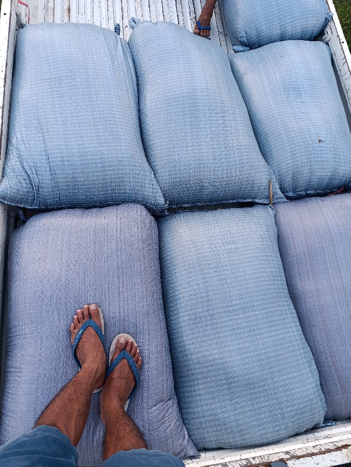 The image shows a truck bed filled with large blue and gray sacks. The sacks are stacked in two rows of three, with the top row slightly offset from the bottom. A person's legs are visible in the lower left corner of the image, with the right foot resting on one of the sacks and the left foot wearing a blue flip-flop. The scene is likely in a rural area near Arjasa, Indonesia, as the coordinates provided (latitude: -7.775, longitude: 114.134) place it in the middle of the island of Java. The surrounding environment is not visible in the image.