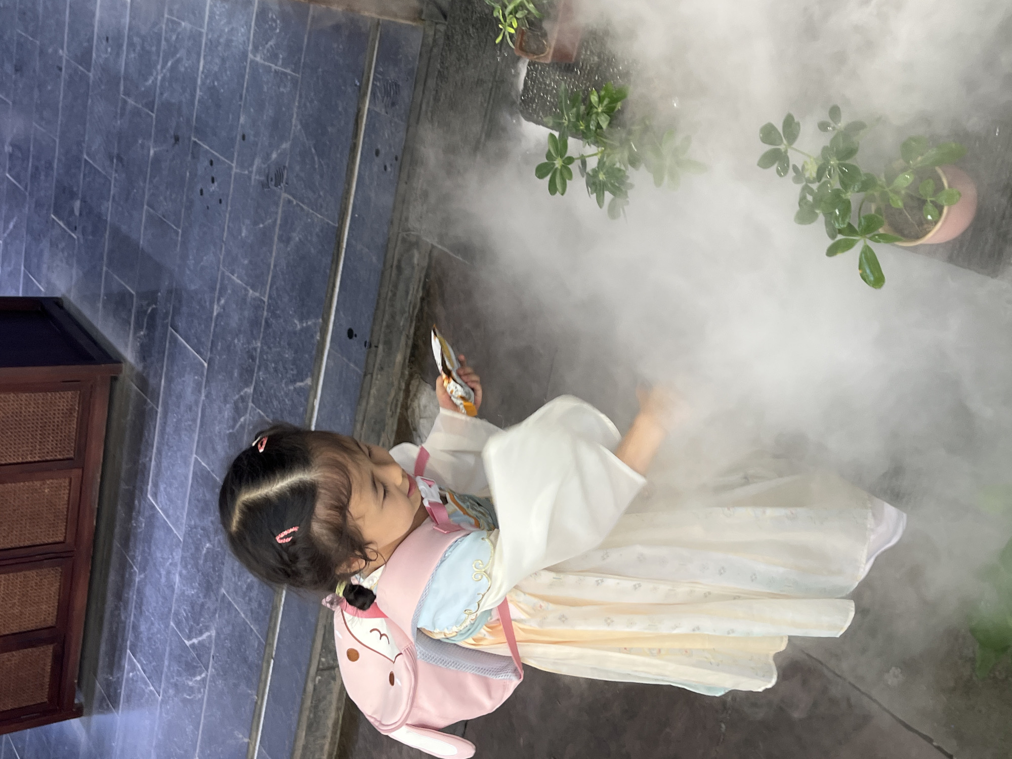 The image shows a young girl wearing a traditional Chinese dress and a pink bunny backpack. She is standing outdoors, surrounded by a dense fog or mist. The girl is holding a small object, possibly a snack, in her right hand and looking down at it. In the foreground, there are several potted plants with green leaves, partially obscured by the mist. The ground is paved with dark, rectangular tiles. In the background, a wooden cabinet or piece of furniture is visible. The scene suggests a garden or outdoor recreational area, possibly in the Yuzhong District of China, given the cultural attire and architectural style. The mist creates a somewhat ethereal atmosphere.