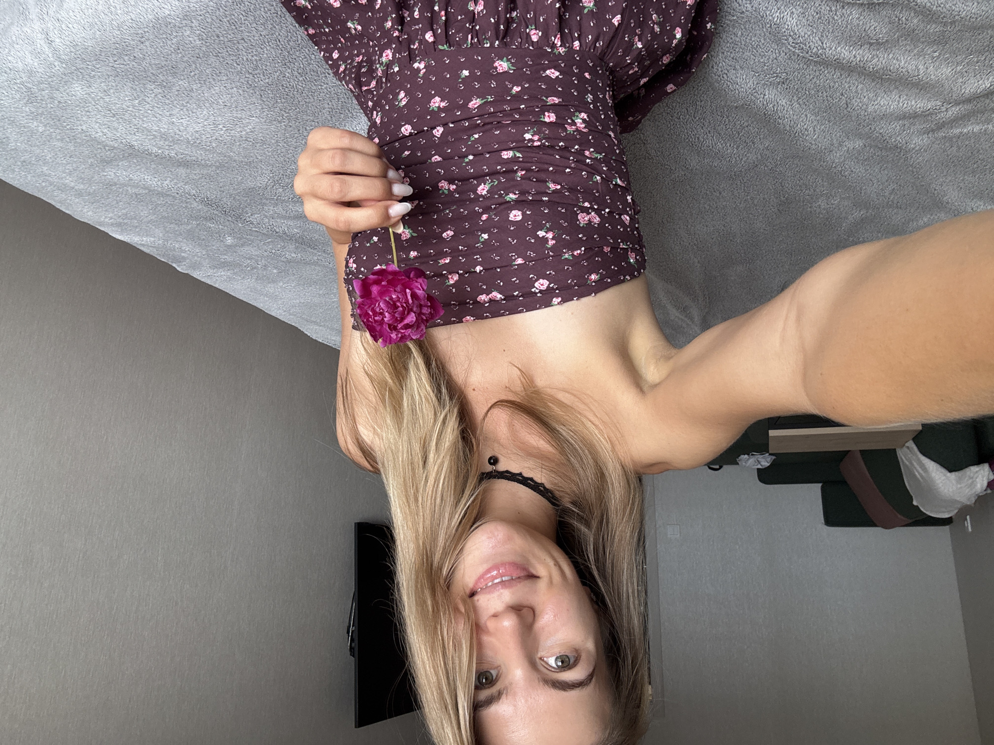 A young woman with blonde hair and green eyes is sitting on a bed in a room. She is wearing a strapless floral-print dress and a black choker necklace. She is holding a single pink flower in her right hand and smiling at the camera. The room is sparsely furnished, with a sofa and a television visible in the background. The overall impression is one of casual intimacy.