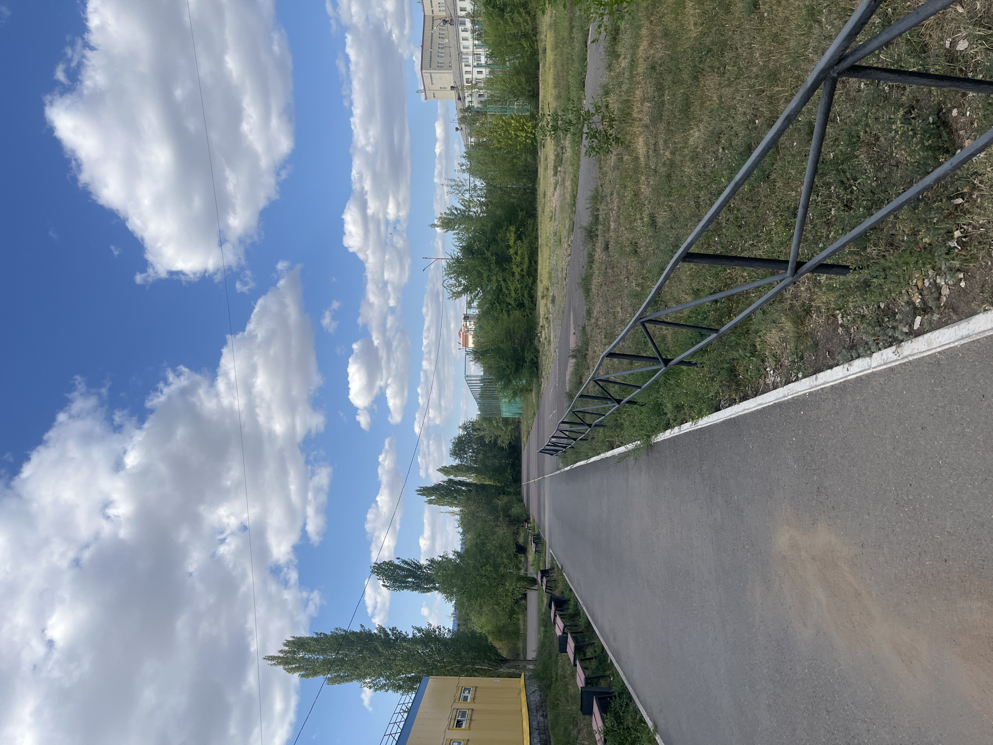 The image depicts a paved pathway running through an outdoor area with a green space on the right and a yellow building on the left. The pathway is bordered by a black metal fence and a white curb. Several benches are visible along the pathway to the left, near the yellow building. Tall, slender trees line the pathway, and in the background, there are more trees, a sports court with green fencing, and residential buildings. The sky is blue with scattered white clouds. This scene is located in Astana, Kazakhstan.