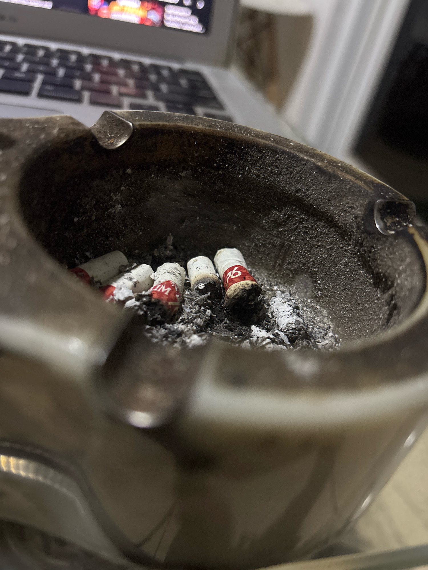 The image shows a close-up of an ashtray filled with several cigarette butts and ash. The ashtray appears to be made of a dark, textured material and is positioned on a surface in front of a laptop keyboard. The cigarette butts have white filters and red bands with some lettering, possibly brand names. The overall scene suggests a casual, indoor environment. No specific location in Bekasi, Indonesia, can be determined from the image.