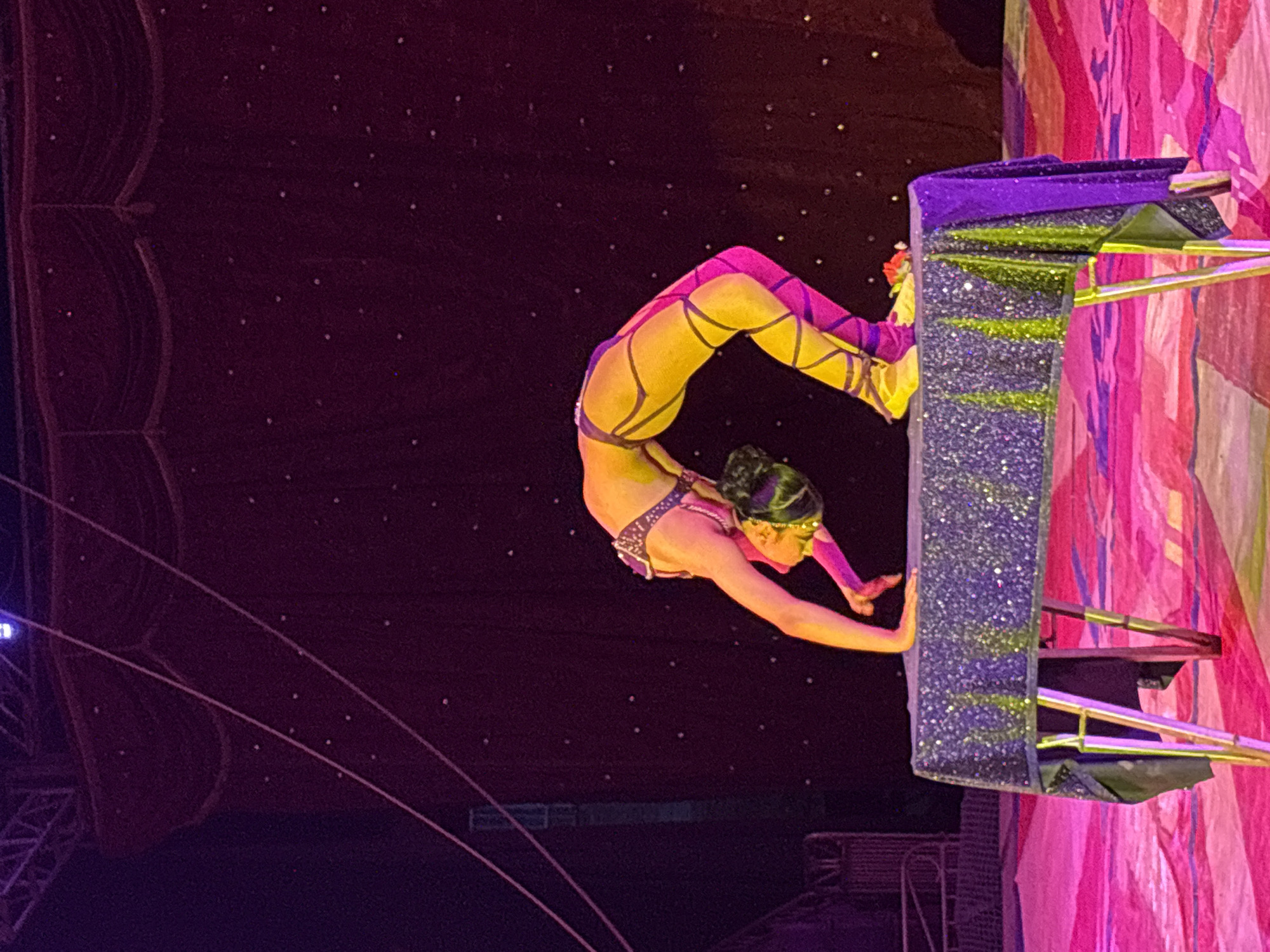 A contortionist performs on a stage during a circus show, possibly in Tejalpa, Mexico. The background appears to be a dark, starry backdrop with red curtains on the left. The contortionist is in a yellow and purple outfit, bent backwards with her hands on a tall structure covered in glitter. The structure has wooden planks attached. The right of the frame has pink and purple fabrics behind the structure.