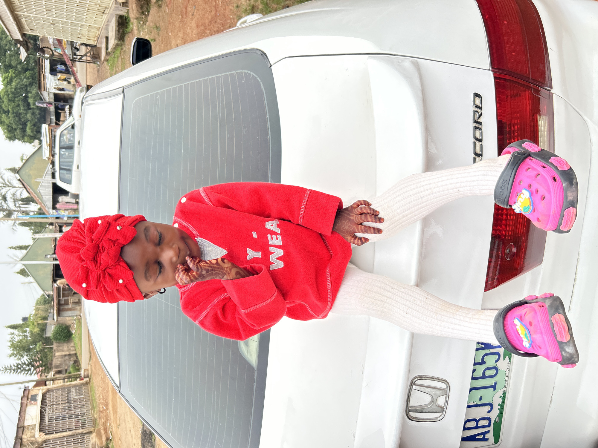 A young child, likely female, is reclining on the trunk of a white Honda Accord. The child is wearing a red, long-sleeved sweater, white tights, and pink Crocs with charms. She is also wearing a red head wrap. The child's hands show signs of henna application. The car has a license plate that reads "ABJ-1651", marked as the Republic of Nigeria. In the background, there are structures and greenery, typical of a populated area. The location appears to be a residential or commercial area in Jos, Nigeria, based on the architectural style of the buildings.