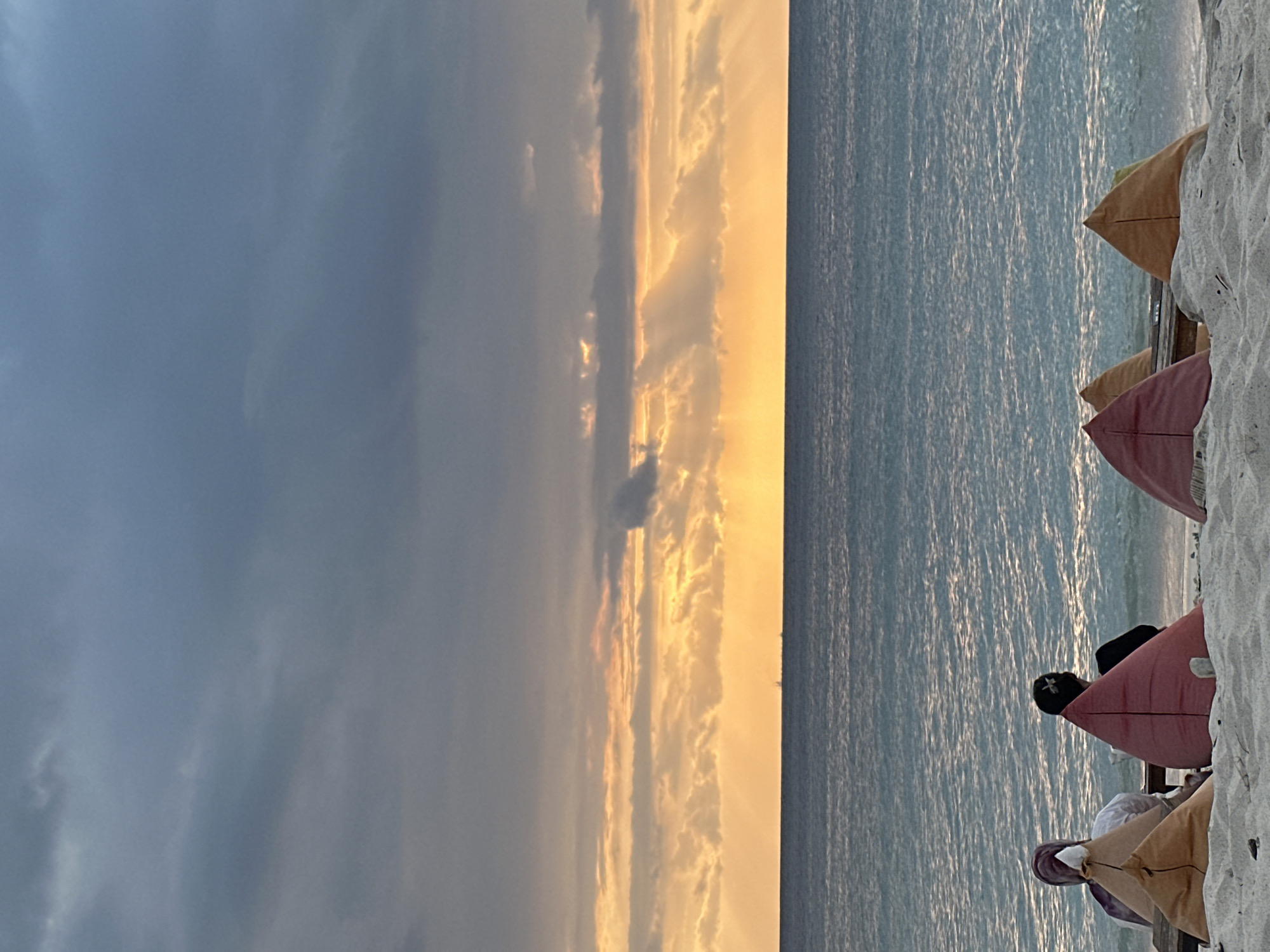 The image shows a beach scene in Unknown, Indonesia, oriented vertically. The sky takes up a significant portion of the left side, displaying a gradient from a dark, overcast gray to a bright golden hue closer to the center. Clouds are scattered across the sky. To the right of the sky is the ocean, with gentle ripples reflecting the light from the sky. In the lower right corner of the image, there are a few pointed beanbag chairs sitting on the sand. Two people are reclined in the beanbag chairs closest to the viewer. The sand is white and textured.