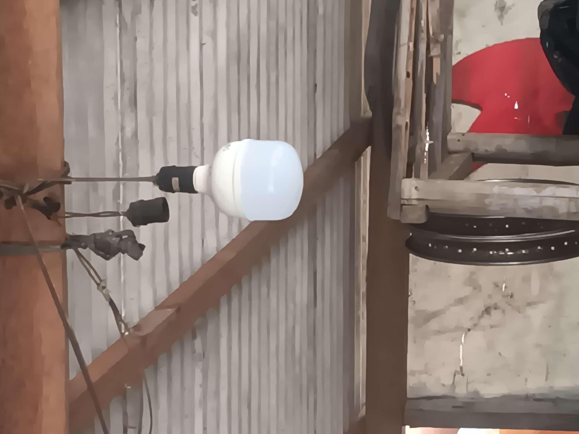 The image depicts an indoor scene, possibly a workshop or storage area, characterized by exposed wooden beams and a corrugated metal ceiling. A prominent object is a large, white LED light bulb hanging from a wire, illuminated from above by a wooden beam. In the background, partially obscured, are various items including what appears to be a bicycle wheel and a large red abstract shape on a wall. The overall impression is of a utilitarian space, possibly in Tlogo, Indonesia, given the context provided. No people are visible in the image, and the scene is static.