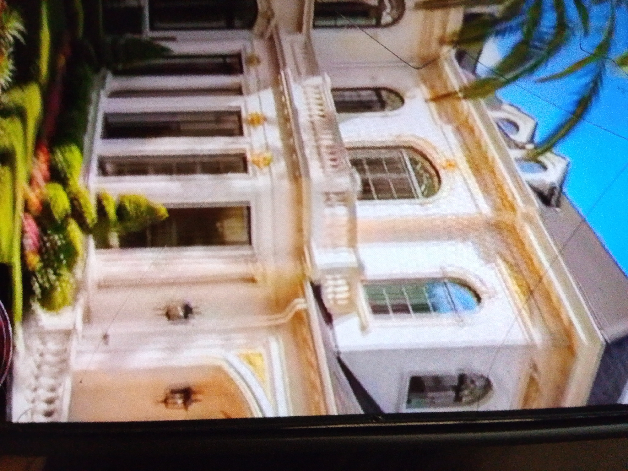The image shows a portion of a large, ornate white building with several windows. The building has multiple levels and a balcony with decorative railings. There are plants with green and pink foliage to the left of the building. Part of a palm tree with a blue sky is visible in the upper right corner. The image appears to be a photograph, possibly taken from a slightly elevated angle, and is of poor quality with visible scratches. The scene is an exterior view of what could be a residential property or a hotel, given its elaborate architecture.