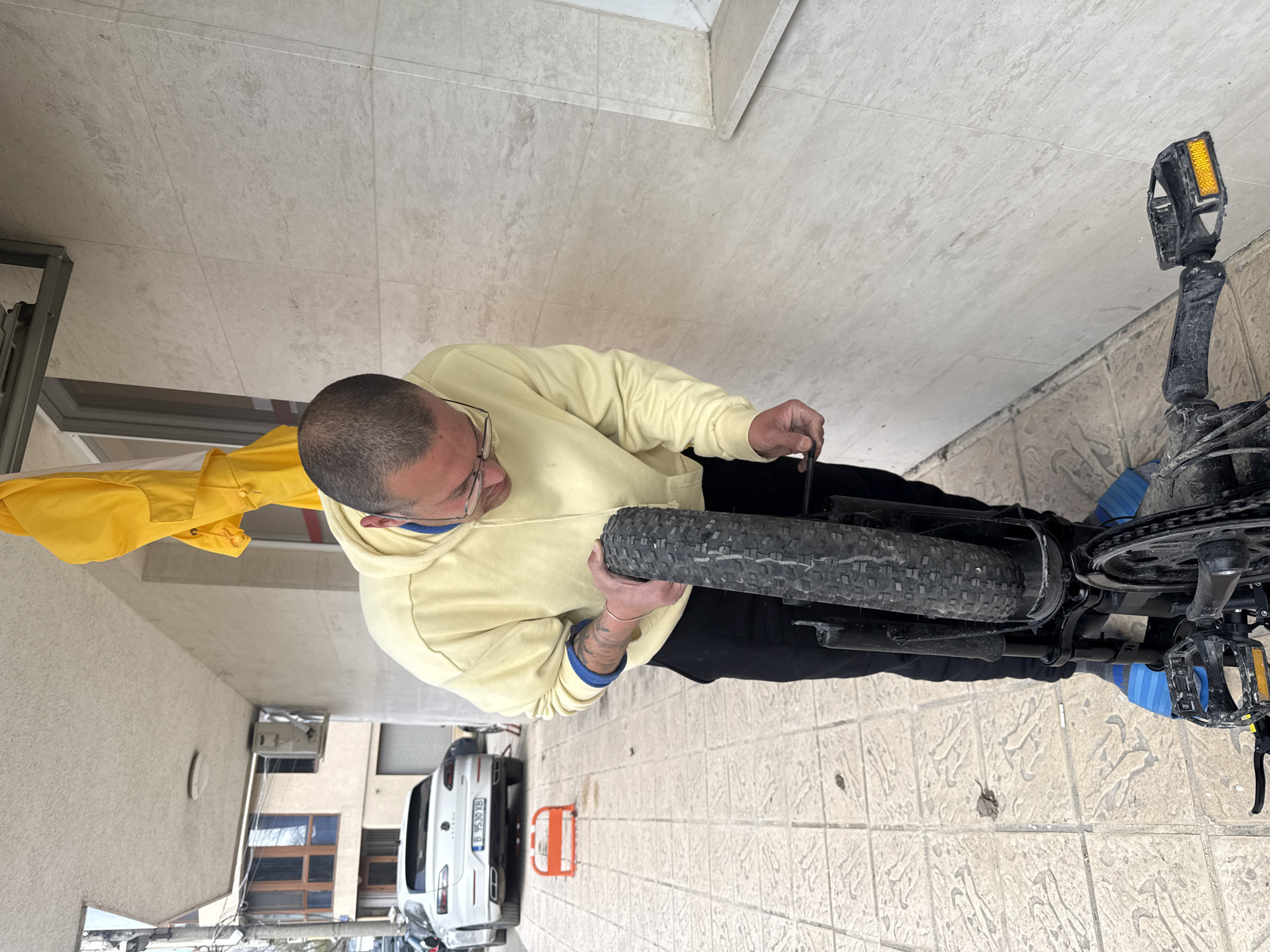 A man in a yellow hoodie and glasses is repairing the rear tire of a black bicycle. The scene takes place outdoors on a tiled surface. A white car with a Bulgarian license plate is parked nearby. The location appears to be an urban residential area. The license plate on the white car indicates a location in Varna, Bulgaria.